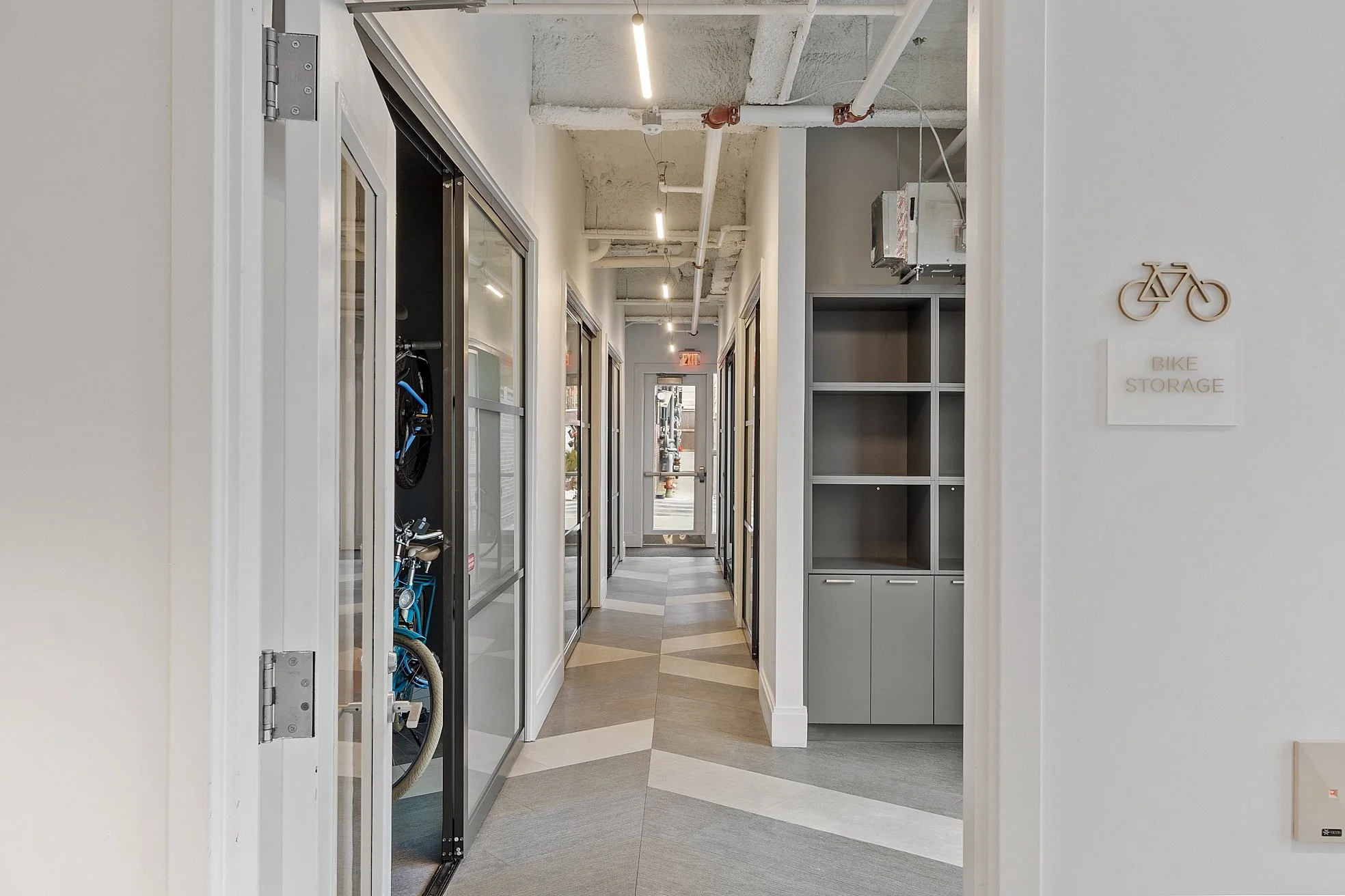 Interior hallway with bike storage area on right side, glass doors on left with bikes inside, geometric patterned flooring, white walls, and ceiling with exposed pipes and lighting fixtures.