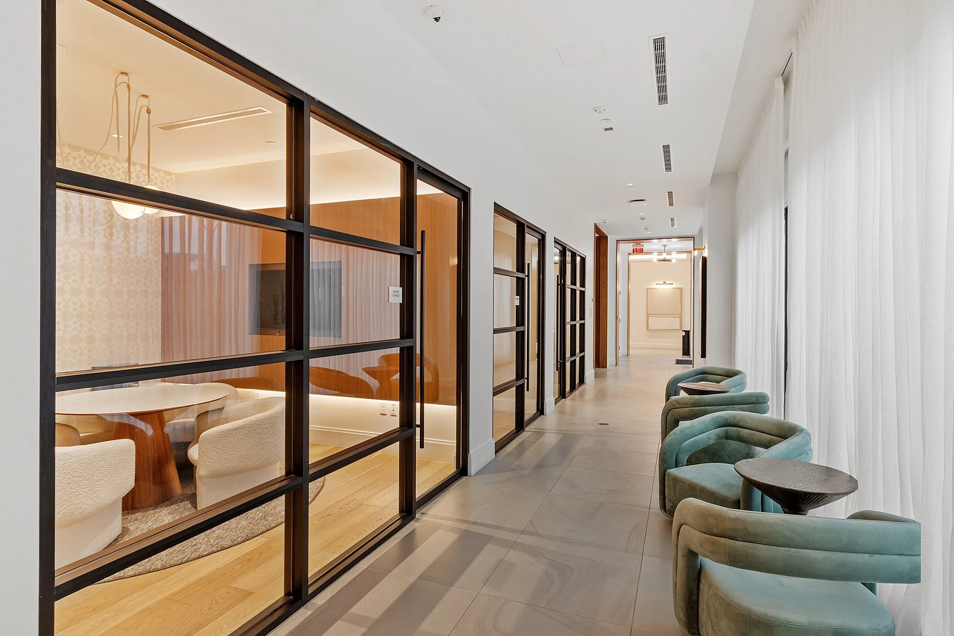 Modern, well-lit hallway with green armchairs and small side tables on the right, and glass-enclosed conference rooms or offices on the left, with a white curtain covering large windows on the right side.