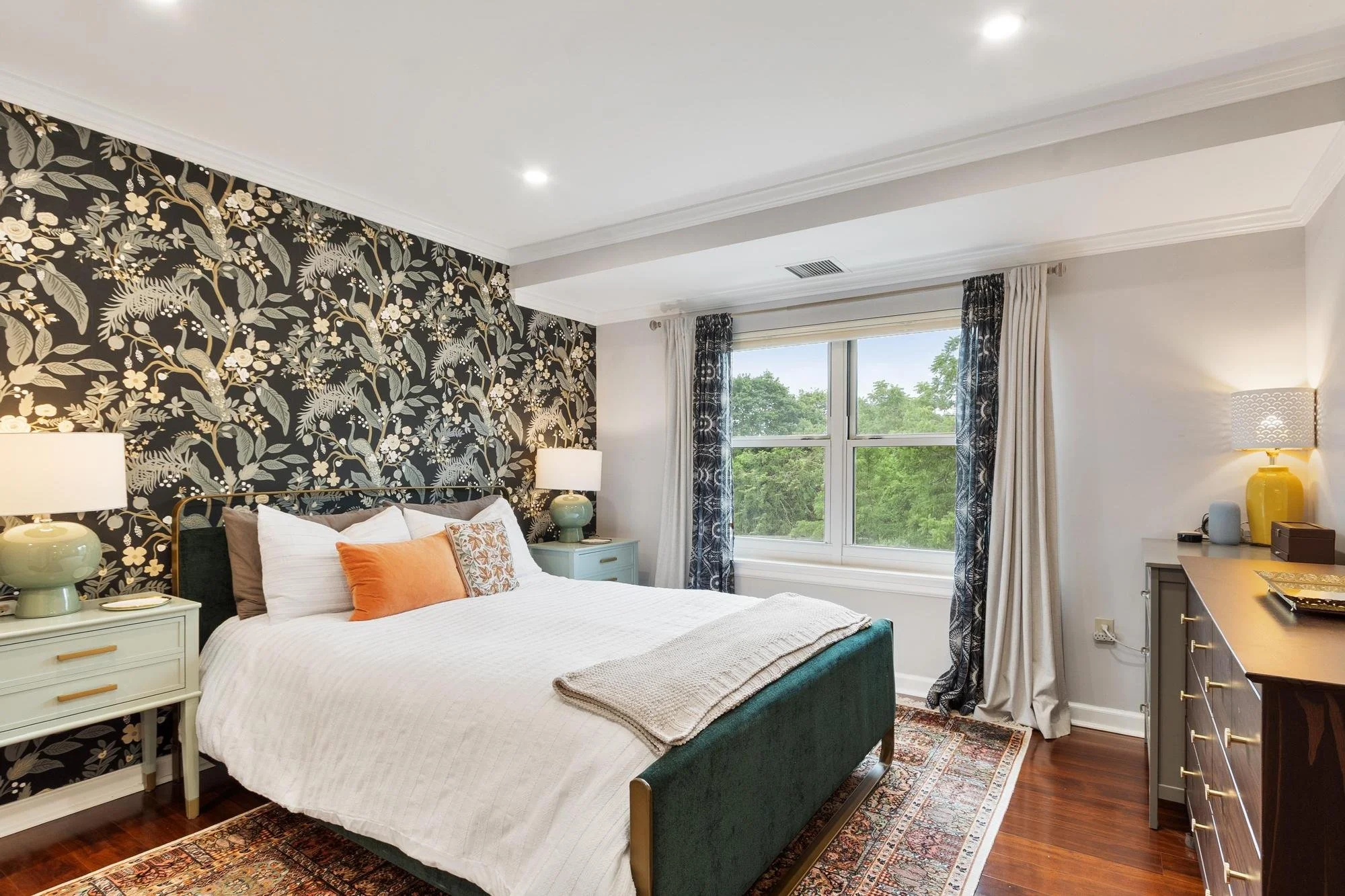 A bedroom with a bed featuring white and orange pillows, a floral black and white accent wall, two matching green lamps on white bedside tables, large window with curtains, desk with yellow lamp, and a patterned area rug.