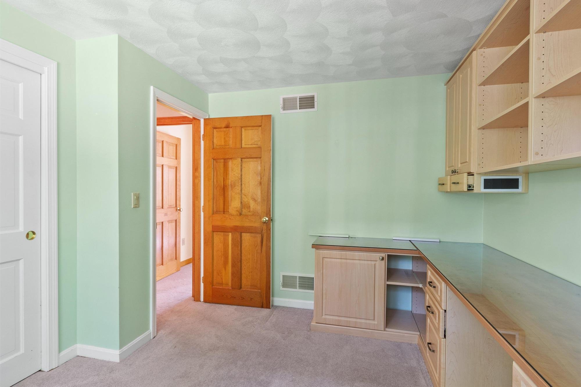 An empty kitchen with light green walls, wooden doors, and unfinished light wood kitchen cabinets. Carpeted floor, small vent on the wall, microwave built into the cabinets.