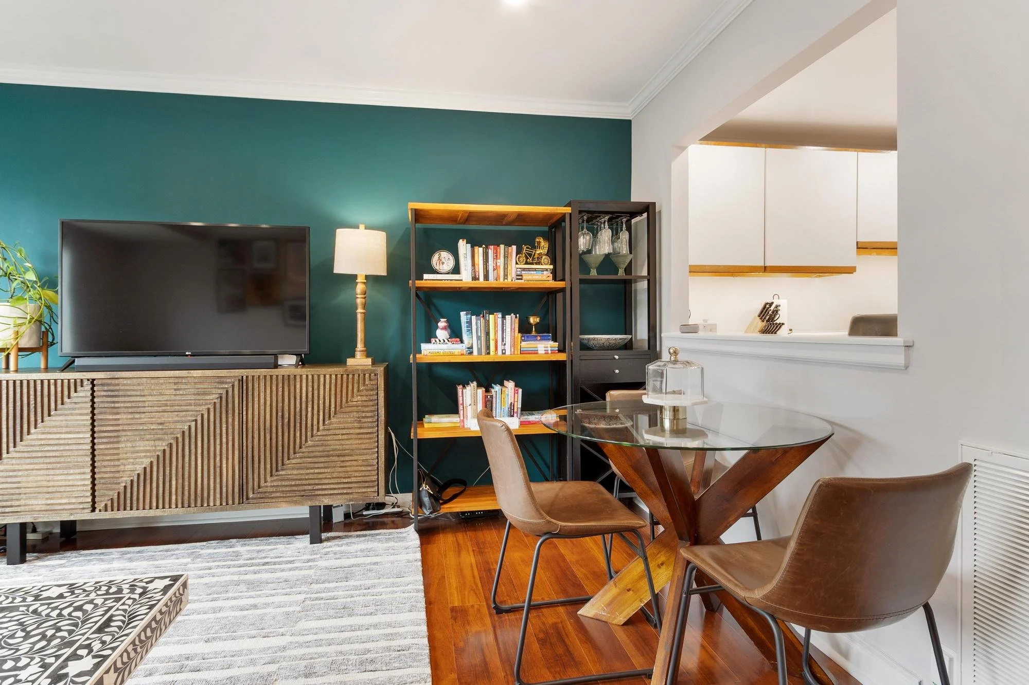 Living room with teal accent wall, a flat-screen TV on a wooden console, a tall floor lamp, a black bookshelf filled with books and decorations, and a glass-top dining table with brown leather chairs. The space leads to a kitchen with white cabinets.