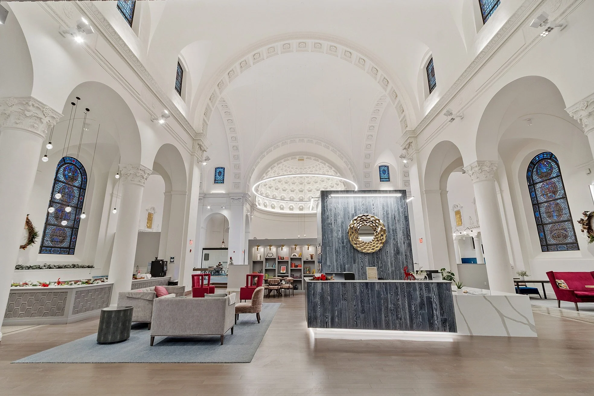 Modern hotel lobby with high arched ceilings, large stained glass windows, and contemporary furniture including chairs and sofas. There is a reception desk with a decorative mirror, a geometric light fixture, and holiday decorations.
