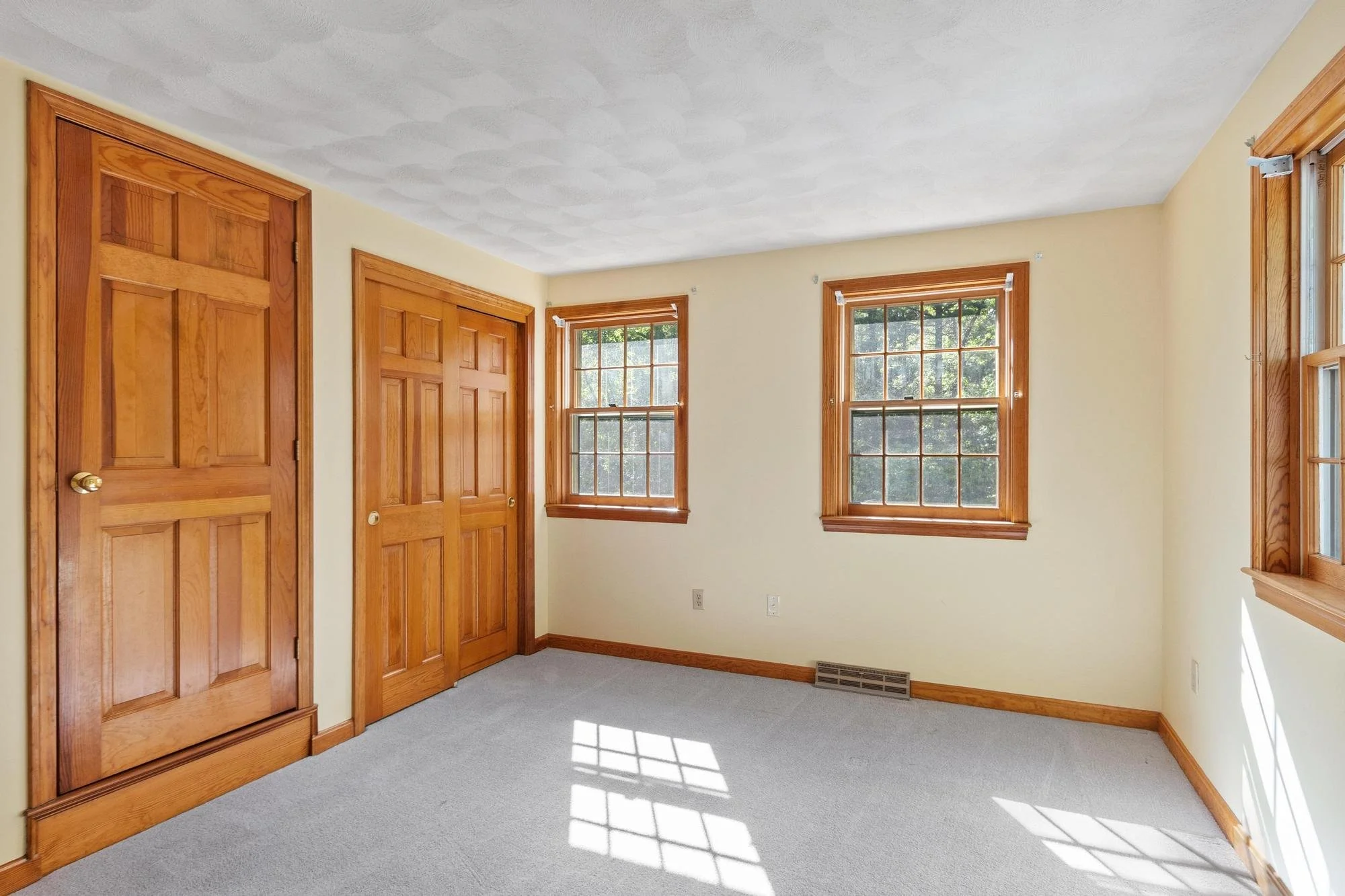 Empty room with beige walls, four wooden framed windows, wooden closet doors, carpeted floor, and baseboard heater.