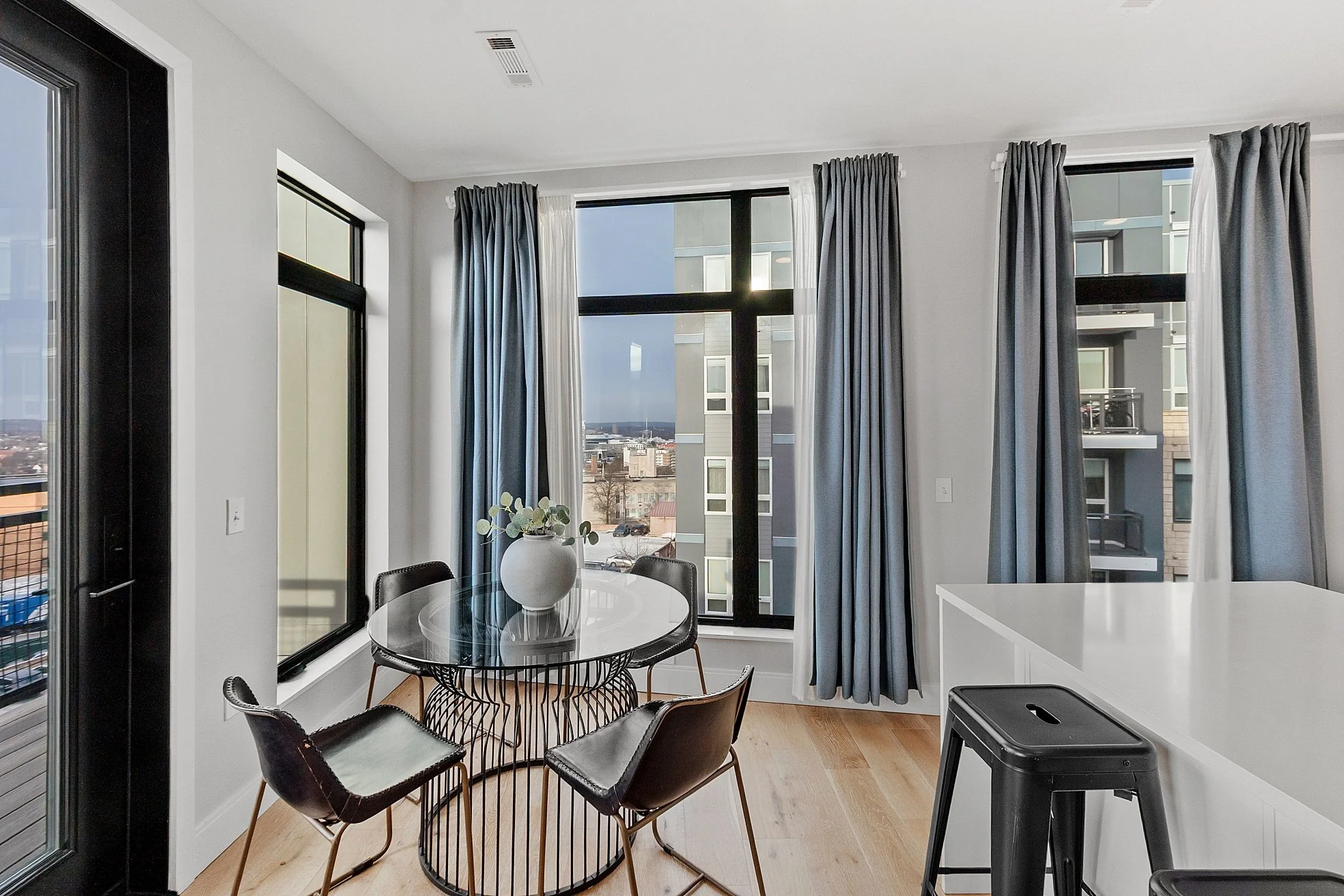 Modern apartment dining area with a round glass table, four chairs, a white vase with greenery, large windows with blue and white curtains, and a white kitchen island with a black stool.