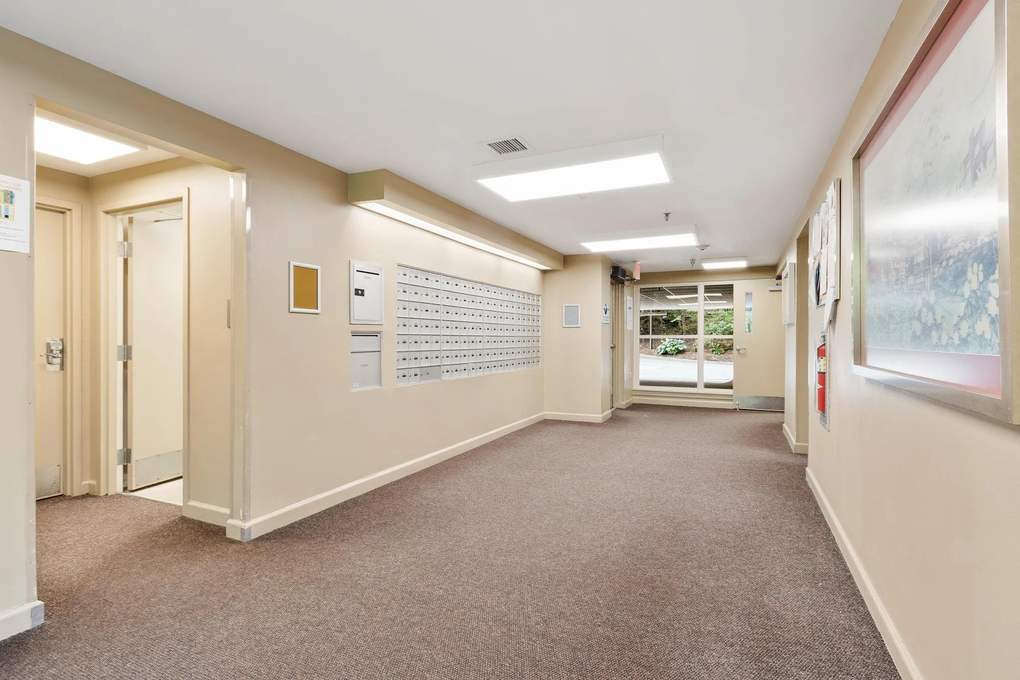 Indoor hallway with mailboxes, bulletin boards, and a fire extinguisher, leading to a glass door and window outside.