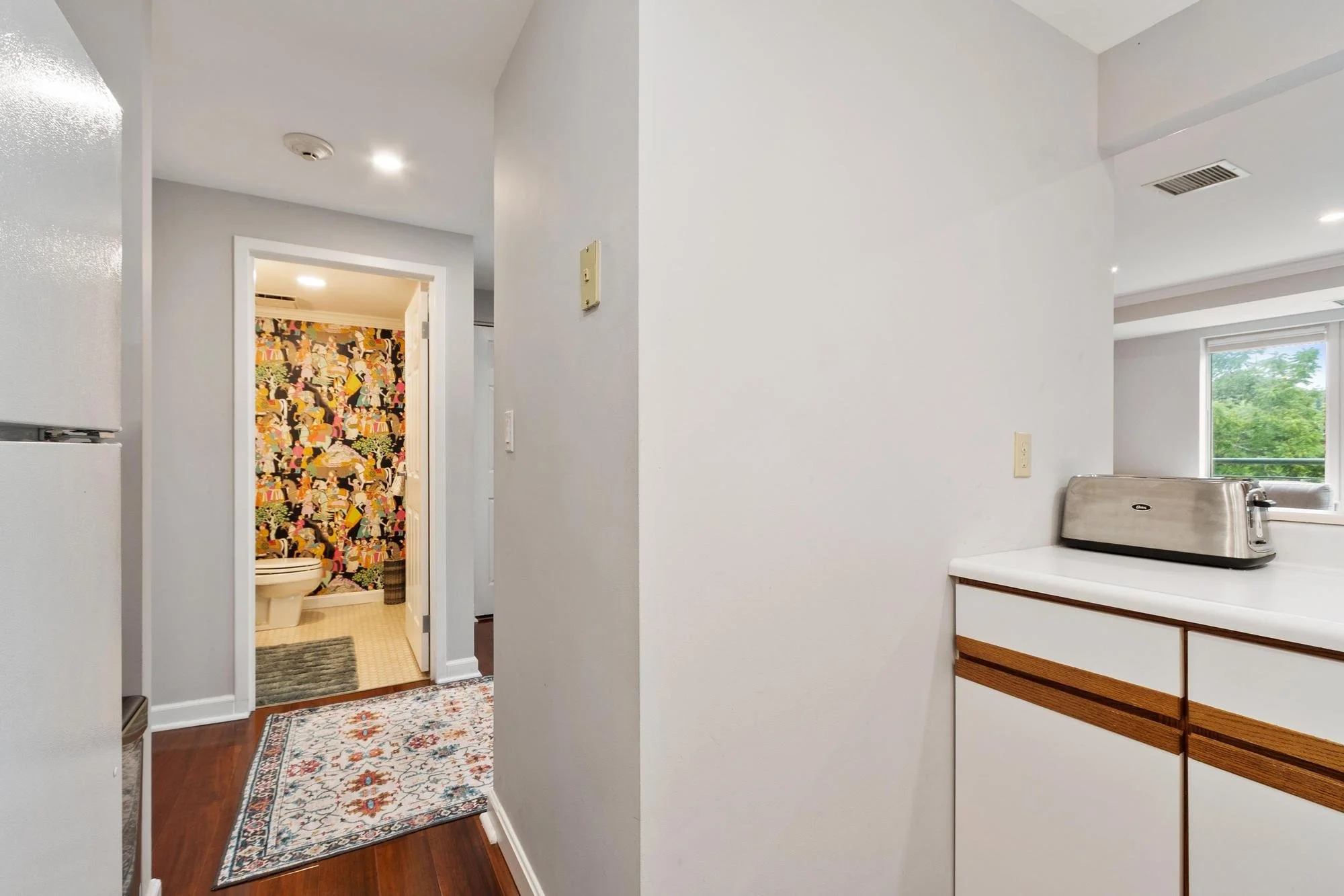 Interior view of a white kitchen with a countertop and toaster, a window with trees outside, a colorful wallpapered bathroom visible through a doorway, and a decorative rug on hardwood floor.