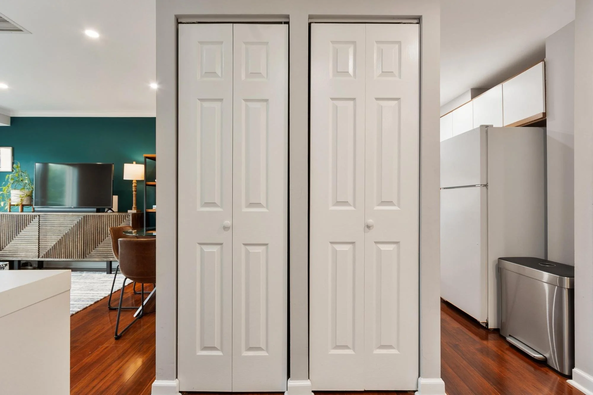 White double-door closet in a modern apartment interior, with wooden floors and partial view of a living room and kitchen.
