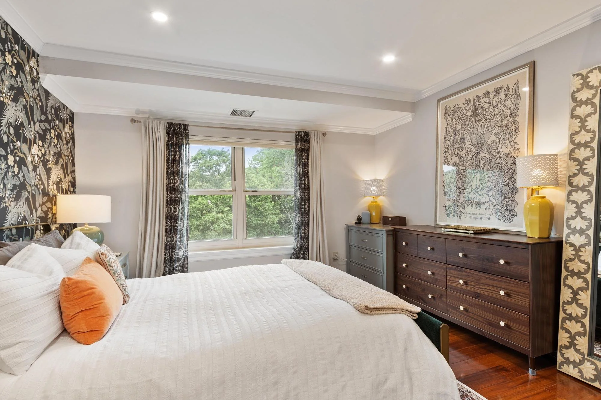 A bedroom with a large window showing green trees outside, a bed with white bedding and orange pillows, a nightstand with a lamp, and a dresser with decorative items and framed art on the wall.
