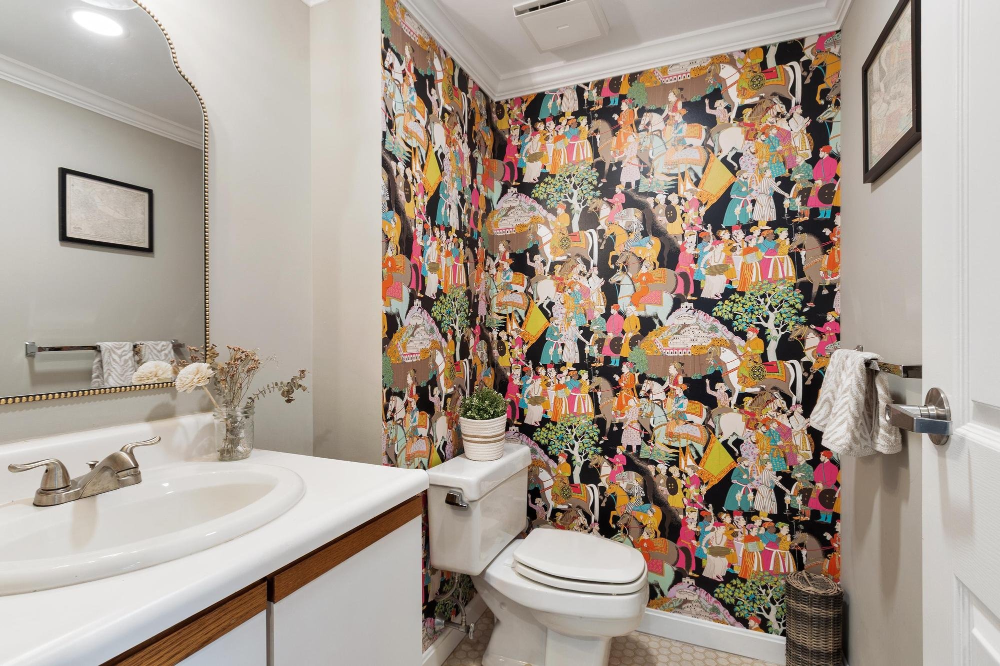 Bathroom with a colorful tapestry shower curtain featuring people in traditional clothing, a white vanity with a sink, a mirror with a bead frame, a framed picture, a white toilet, a basket, and decorative plants.