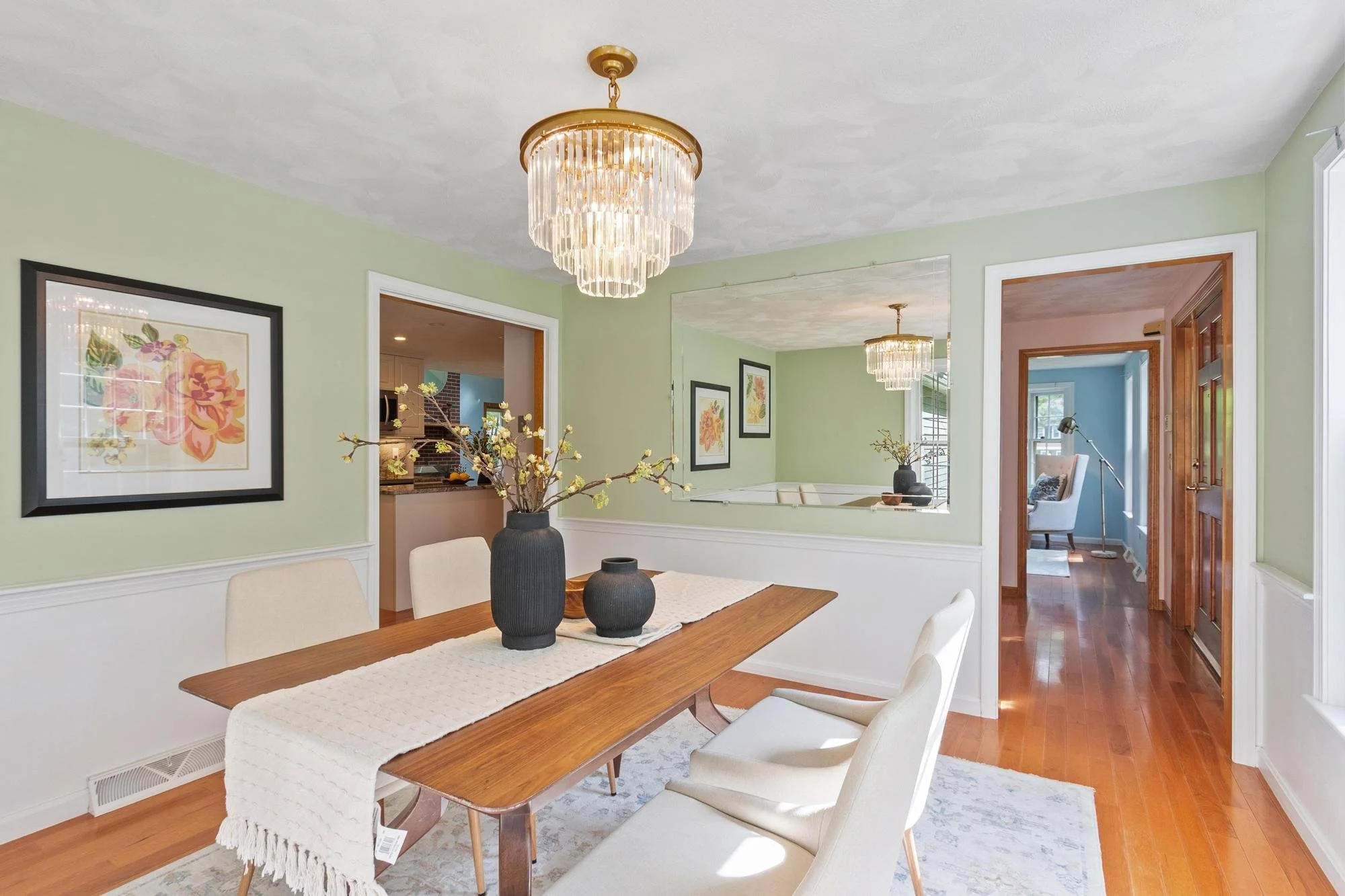 Dining room with wooden table and white chairs, black vases with flowers, framed floral artwork on light green walls, wooden floor, and crystal chandeliers overhead.