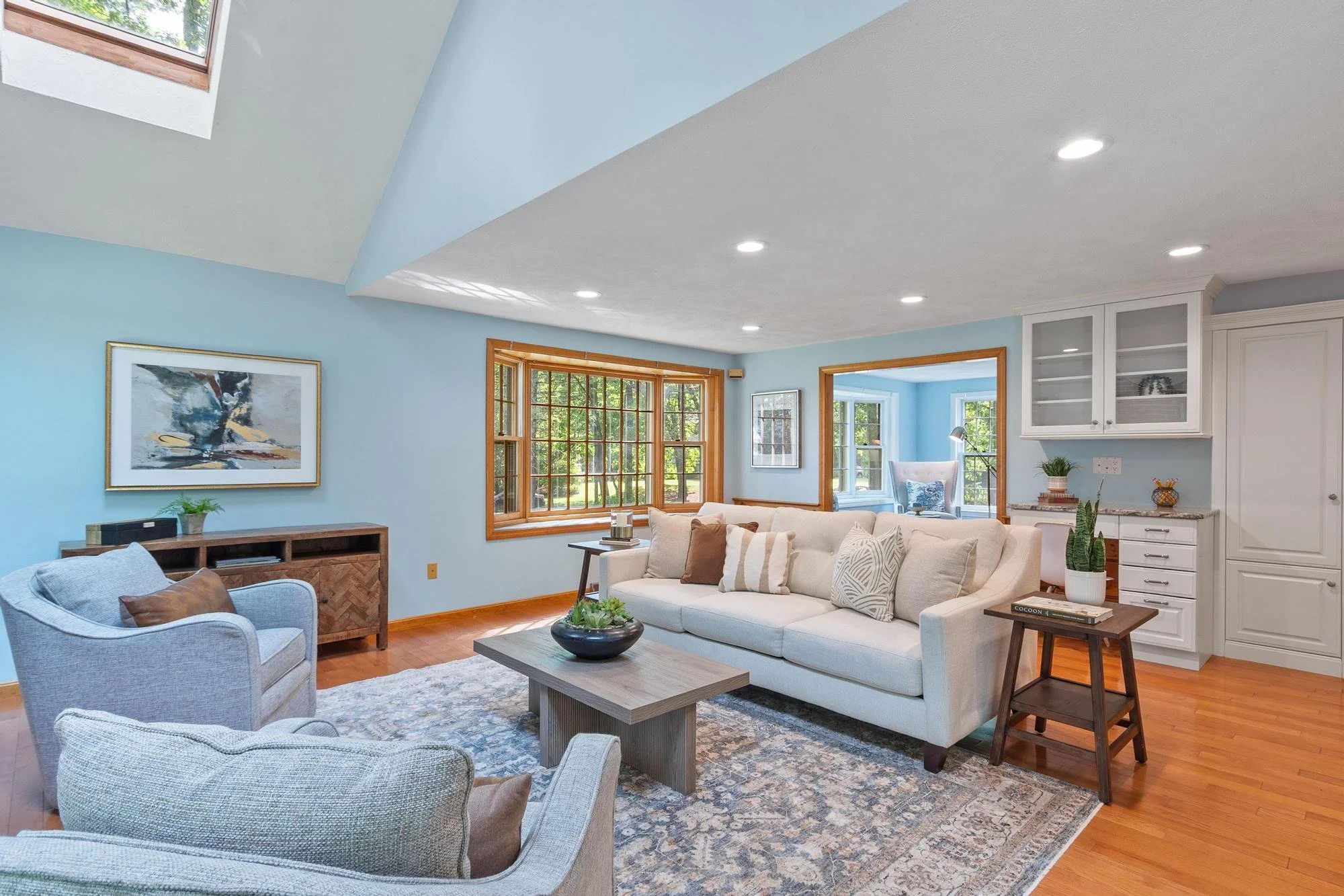 Living room with light blue walls, a white sofa with beige and patterned pillows, two armchairs, a wooden coffee table, windows with wooden trim, artwork on the walls, a potted plant, and recessed ceiling lights.