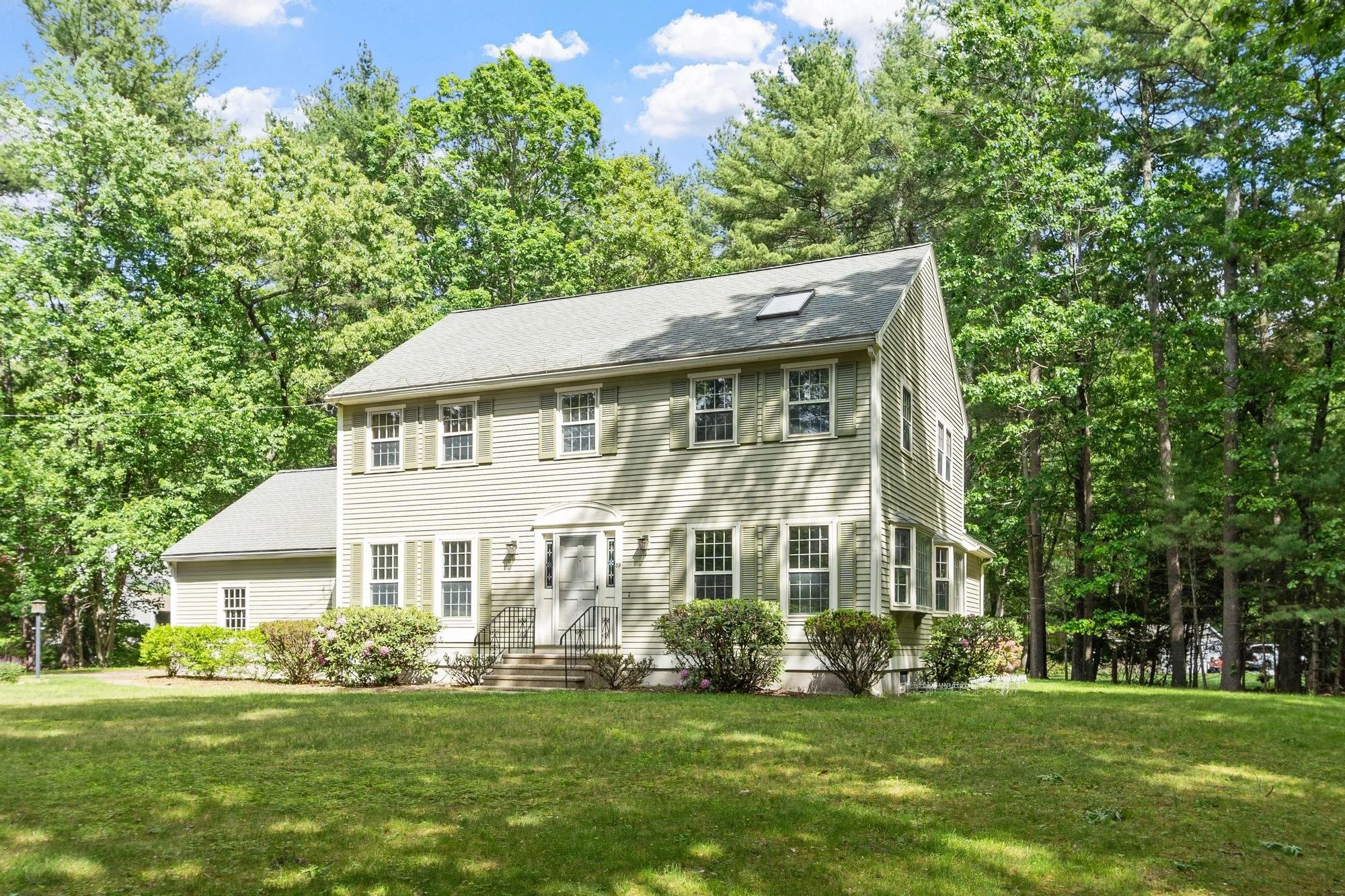 A beige, three-story house with multiple windows and shutters, surrounded by green bushes and trees, under a partly cloudy sky.