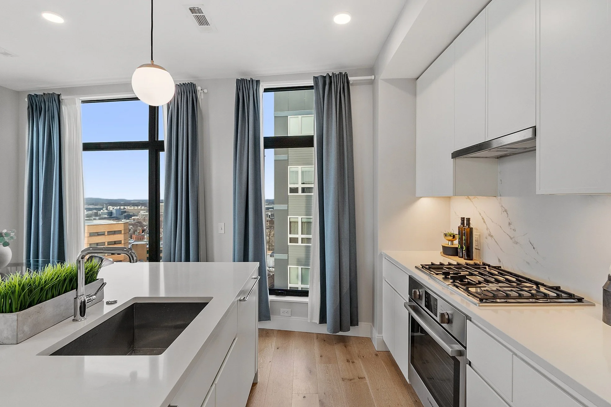 Modern kitchen with white cabinetry, marble backsplash, stainless steel appliances, and large windows with blue curtains overlooking a cityscape.