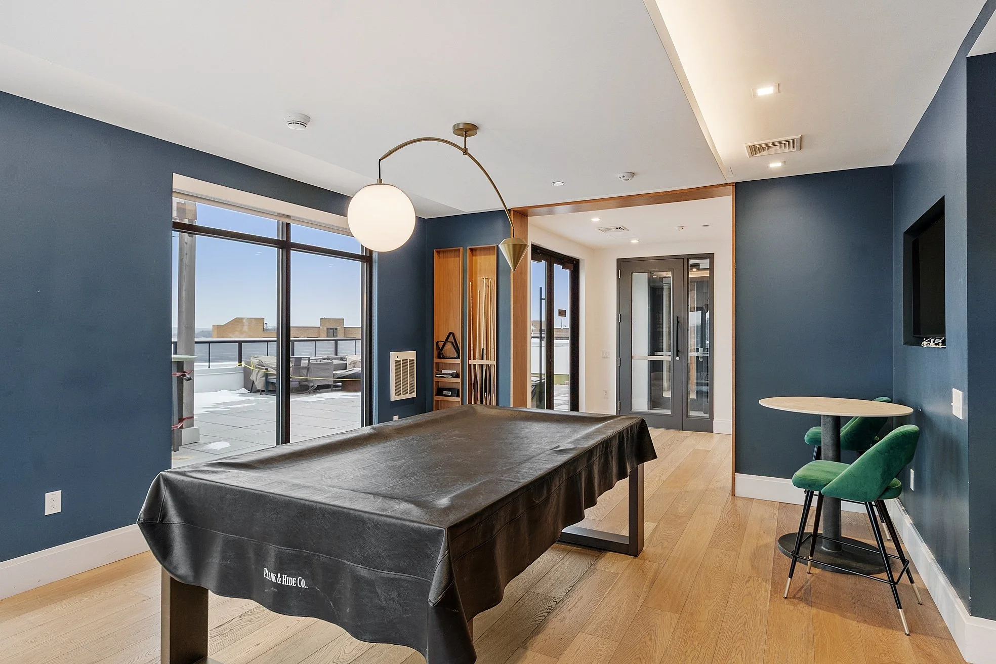 Modern dining or lounge area with blue walls, hardwood floors, large sliding glass doors leading to balcony, black pool table, green velvet bar stools, and small round dining table.