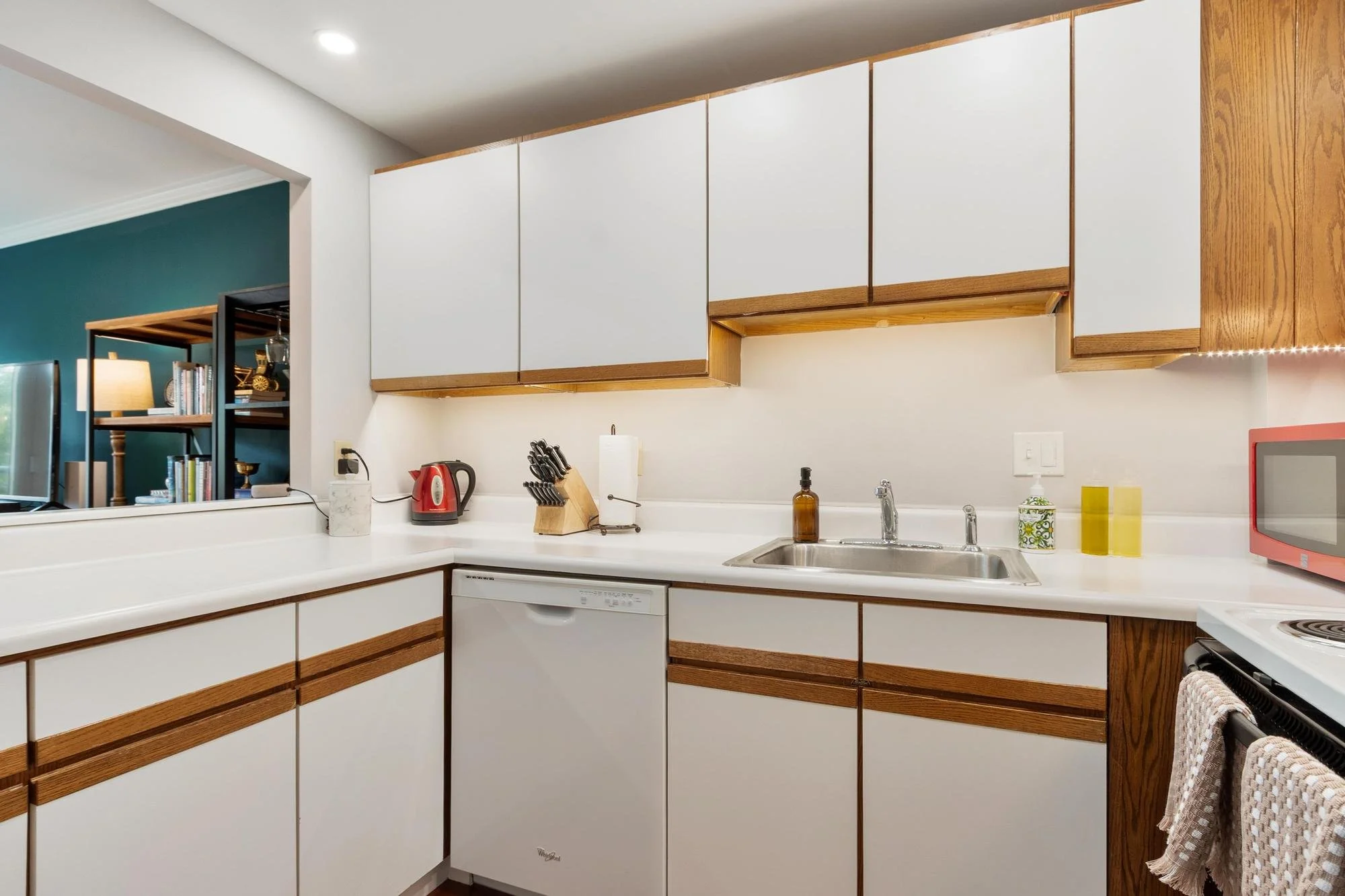 Kitchen with white cabinets and wooden trim, white countertop, stainless steel sink, small appliances, and decorative items.