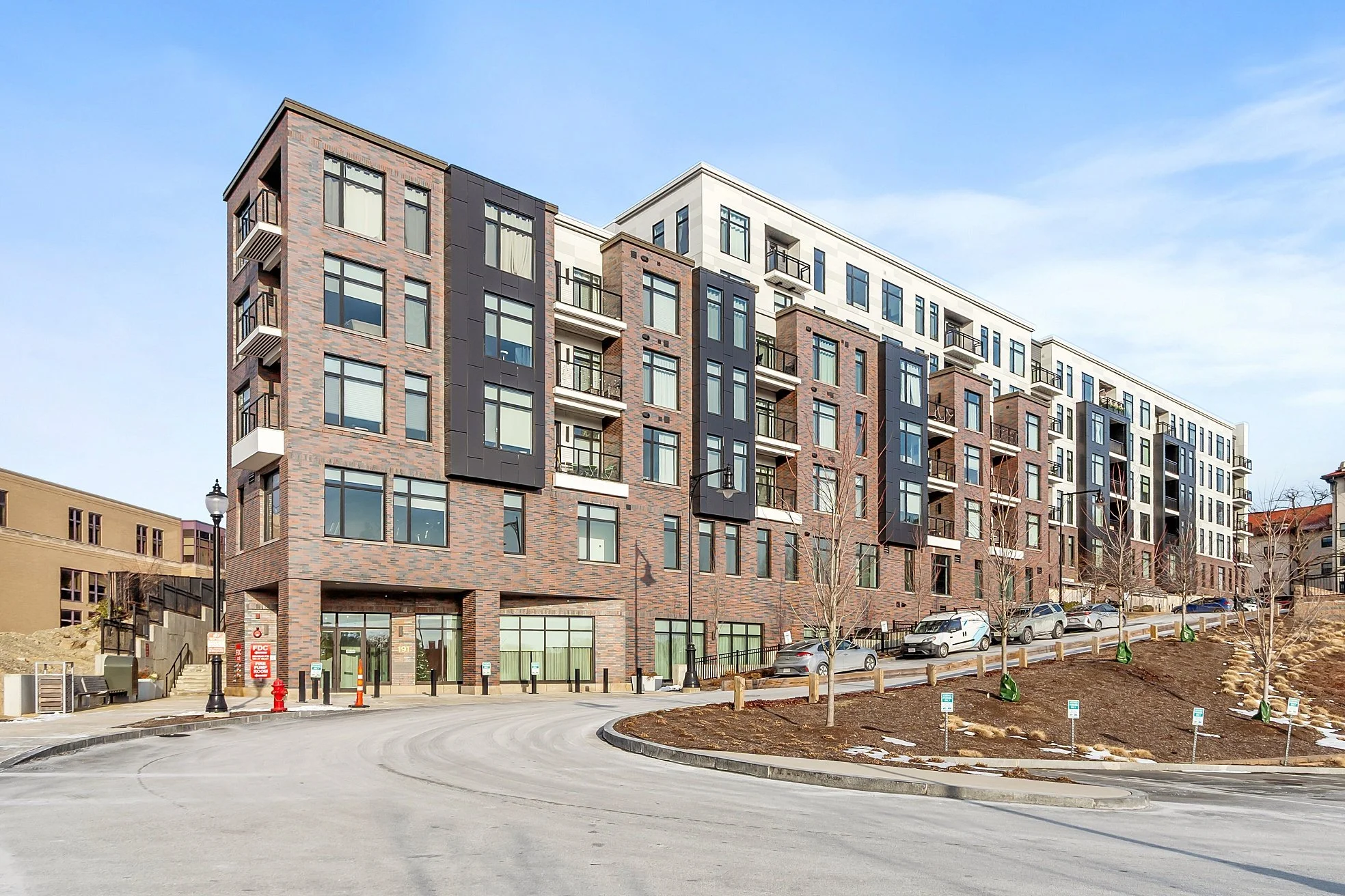 Modern multi-story apartment building with brick and white facade, balconies, and large windows, situated along a curved driveway with parked cars and leafless trees.
