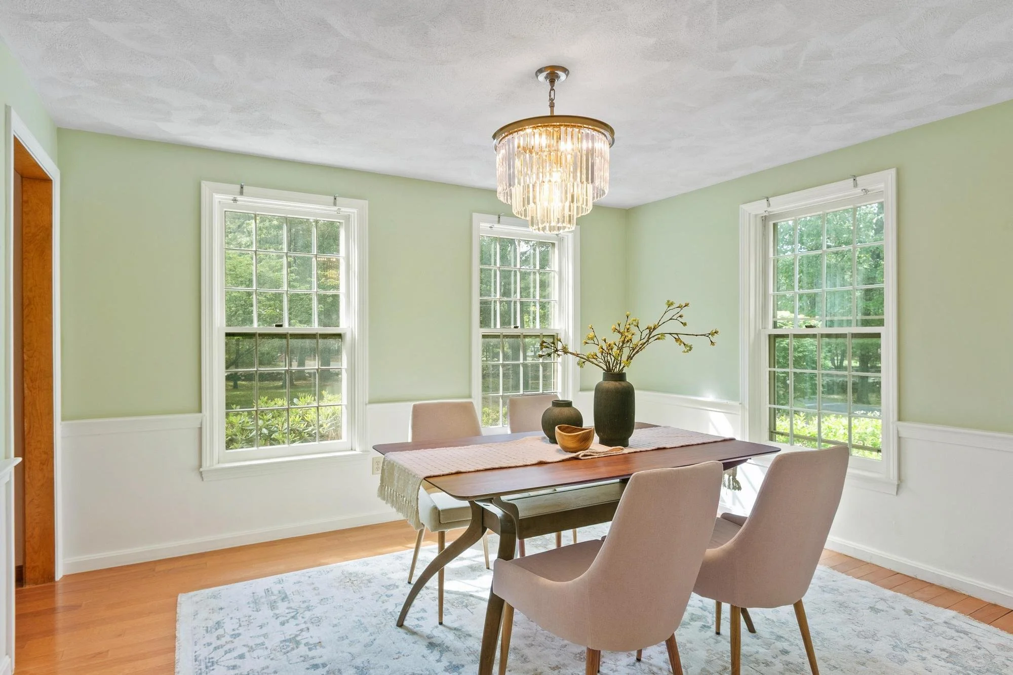 A dining room with three large windows, a wooden dining table with a beige tablecloth, and four beige upholstered chairs. There are two black vases with branches and a wooden bowl on the table. A chandelier hangs from the ceiling. The walls are painted light green, and the floor is hardwood with a patterned rug.