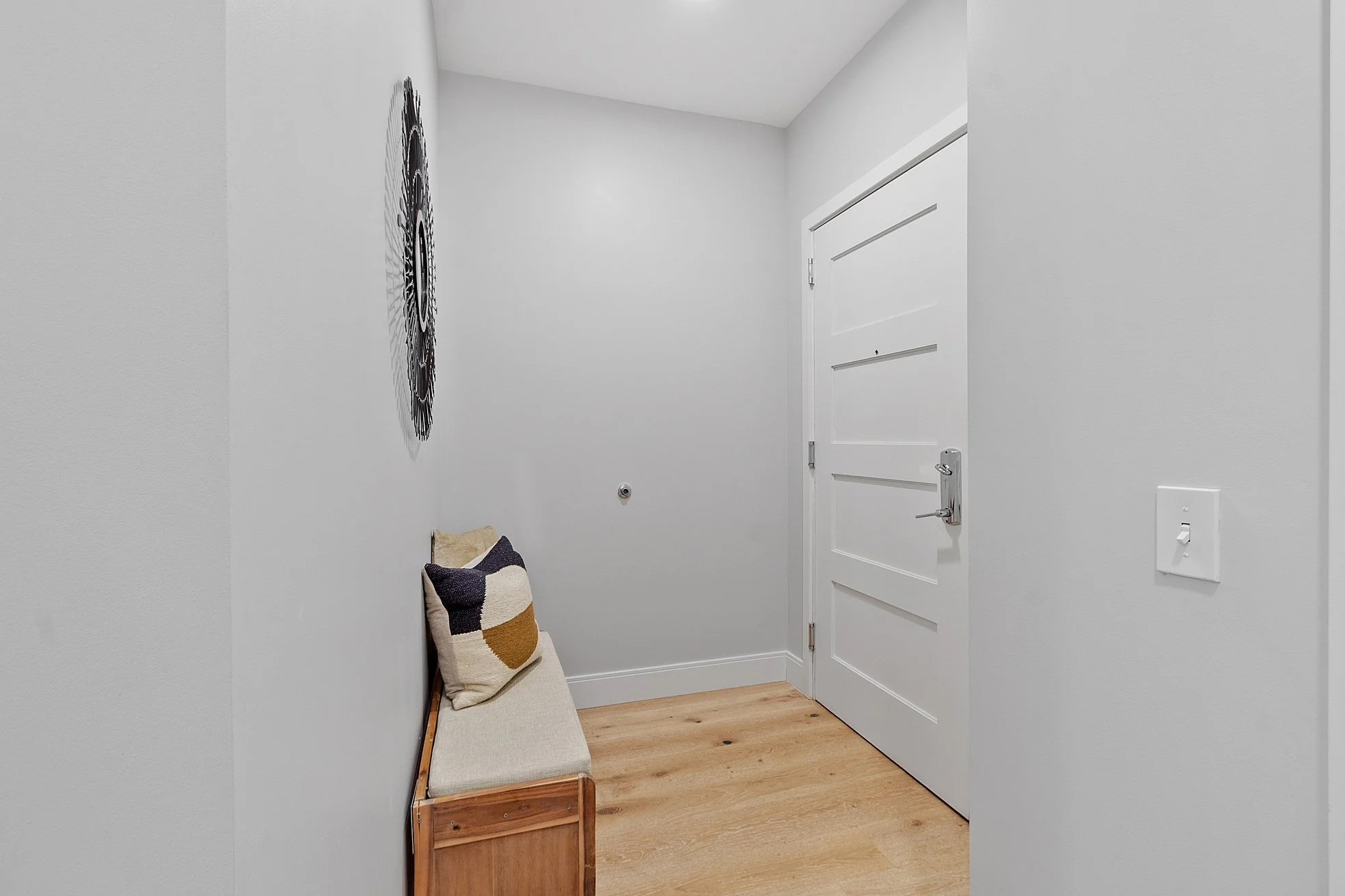 Small entryway with white door, wooden bench with beige cushion and pillows, decorative wall art, hardwood floor.