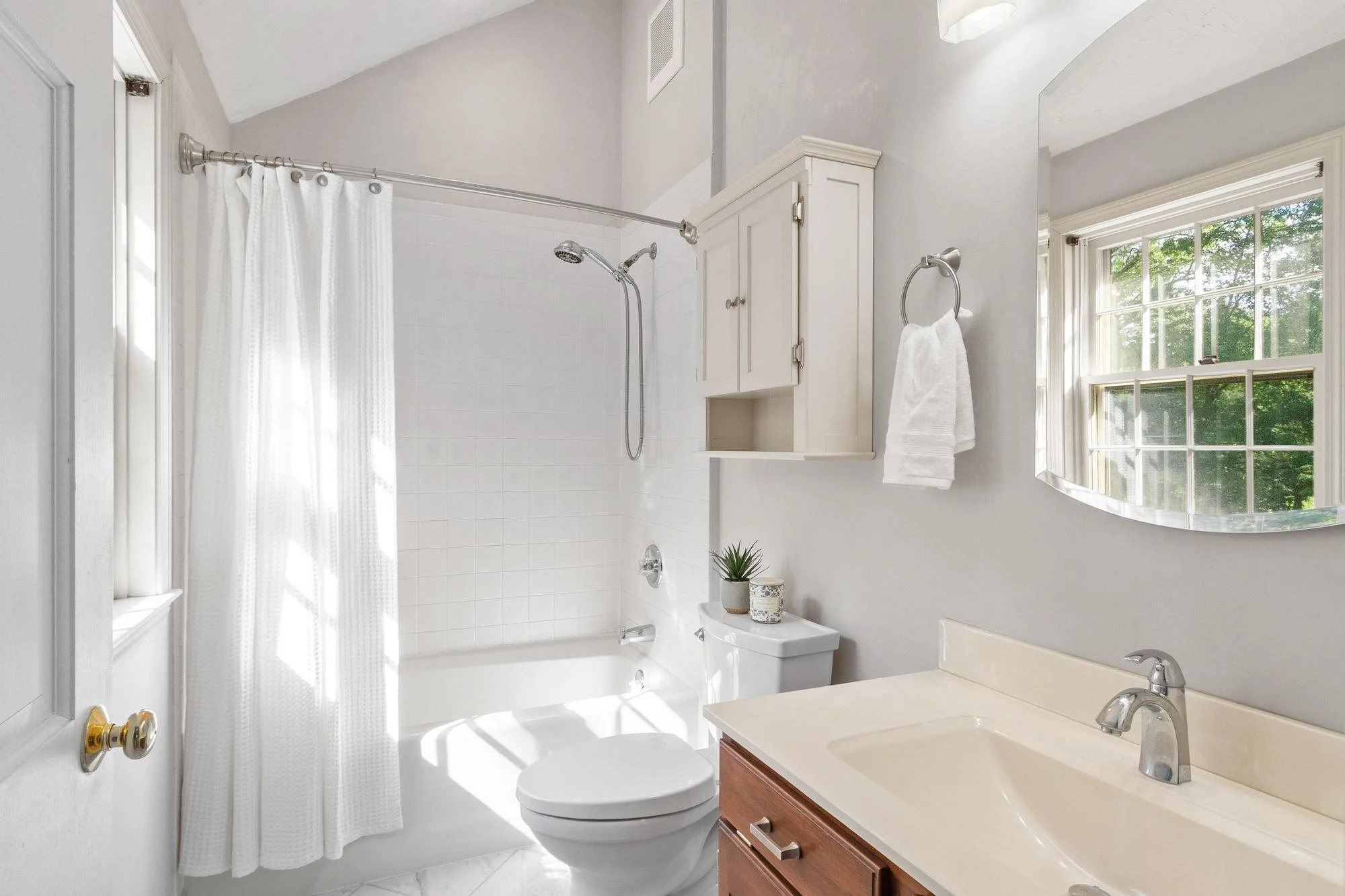 Bright bathroom with white tiles and walls, featuring a shower curtain, window with greenery outside, white built-in cabinets, a toilet, a small plant, and a mirror over the sink.