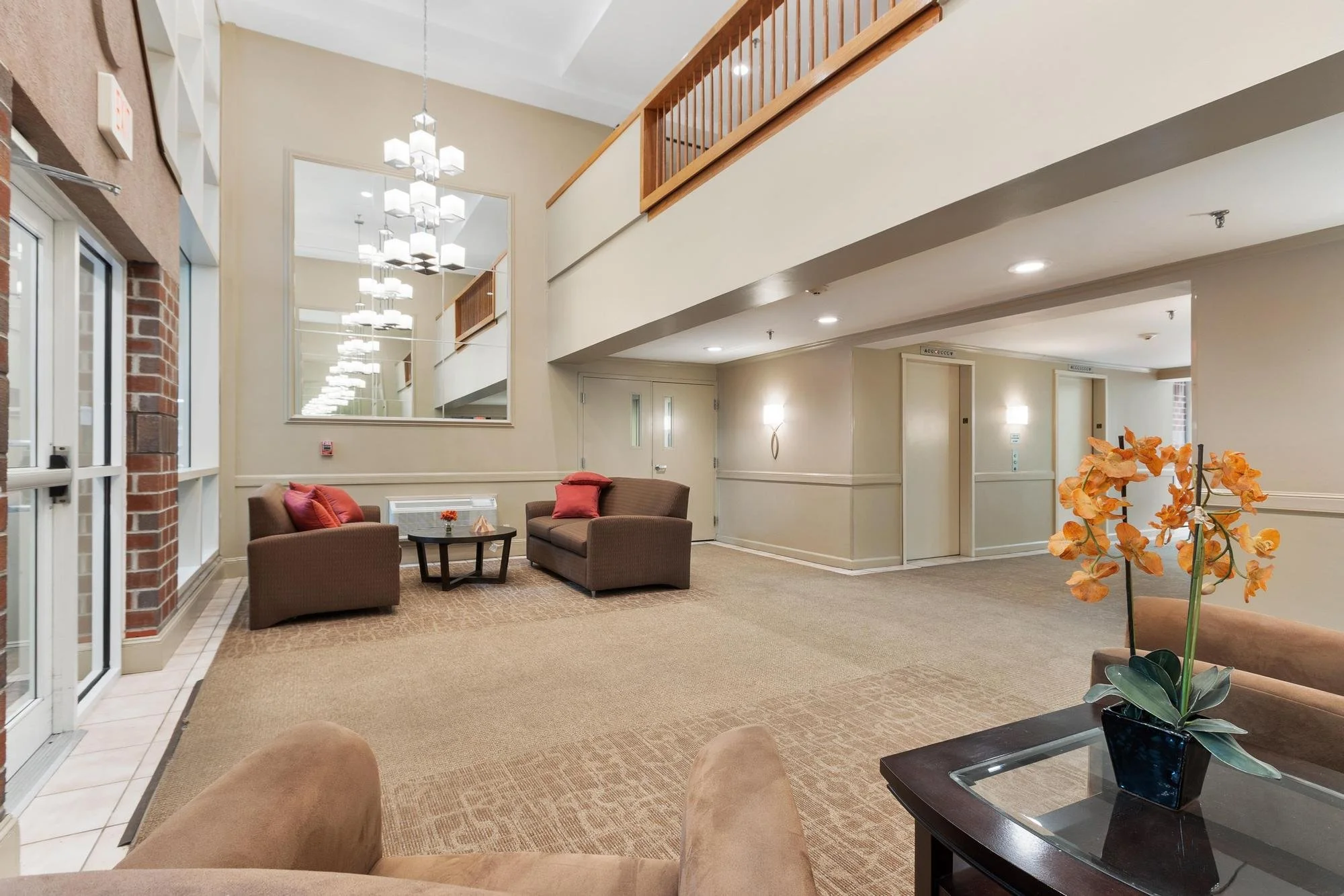 Lobby or waiting area with beige carpet, brown armchairs with red pillows, a small black round table with a flower, wall sconces, a large mirror, a flower arrangement with orange orchids on a glass table, beige walls, and wooden railing on the upper level.