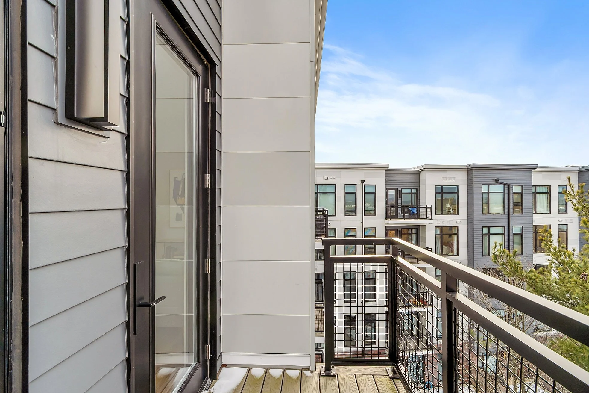 A small balcony with black metal railing, overlooking modern apartment buildings, with a sliding glass door and partial view of the interior.