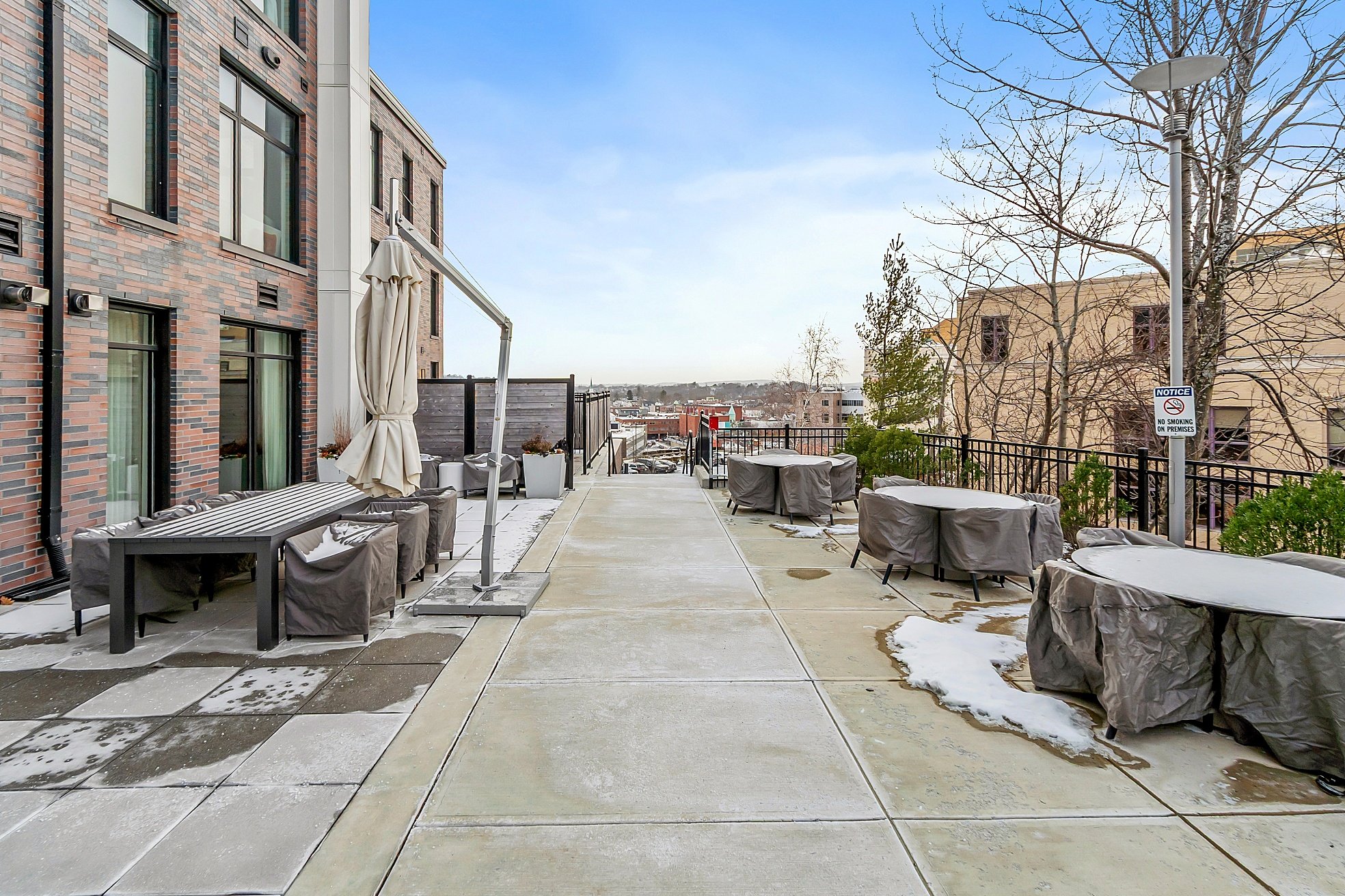 Empty outdoor patio with tables and chairs covered in gray protective covers, some snow patches on the ground, a brick building on the left, leafless trees, a lamp post, and a distant cityscape under a blue sky.