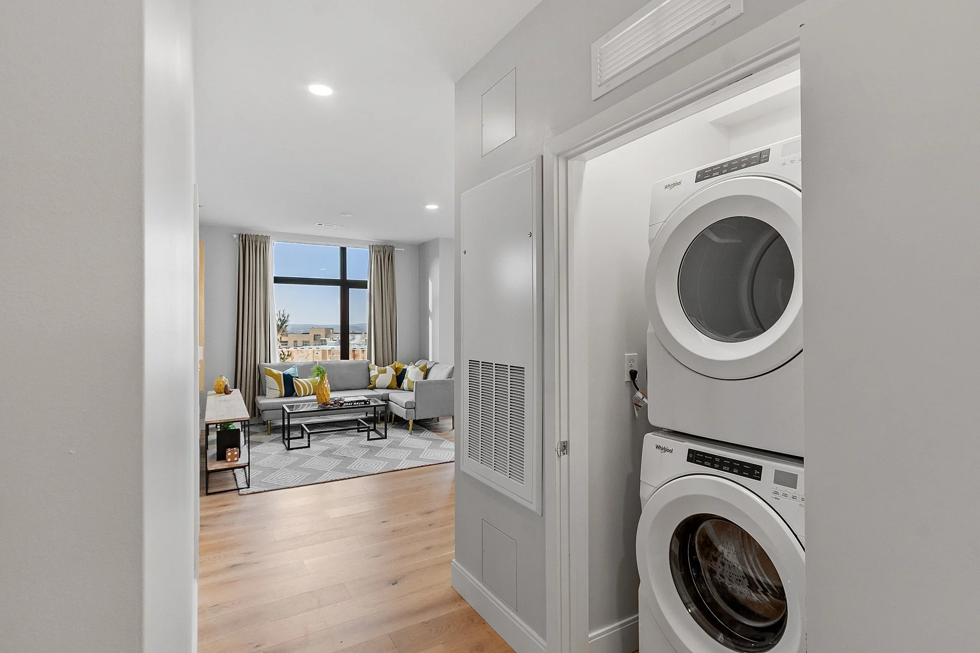 View of a living room with a large window and beige curtains, a gray sofa with colorful pillows, a coffee table, and a wooden side table, with a patterned rug on light hardwood flooring.