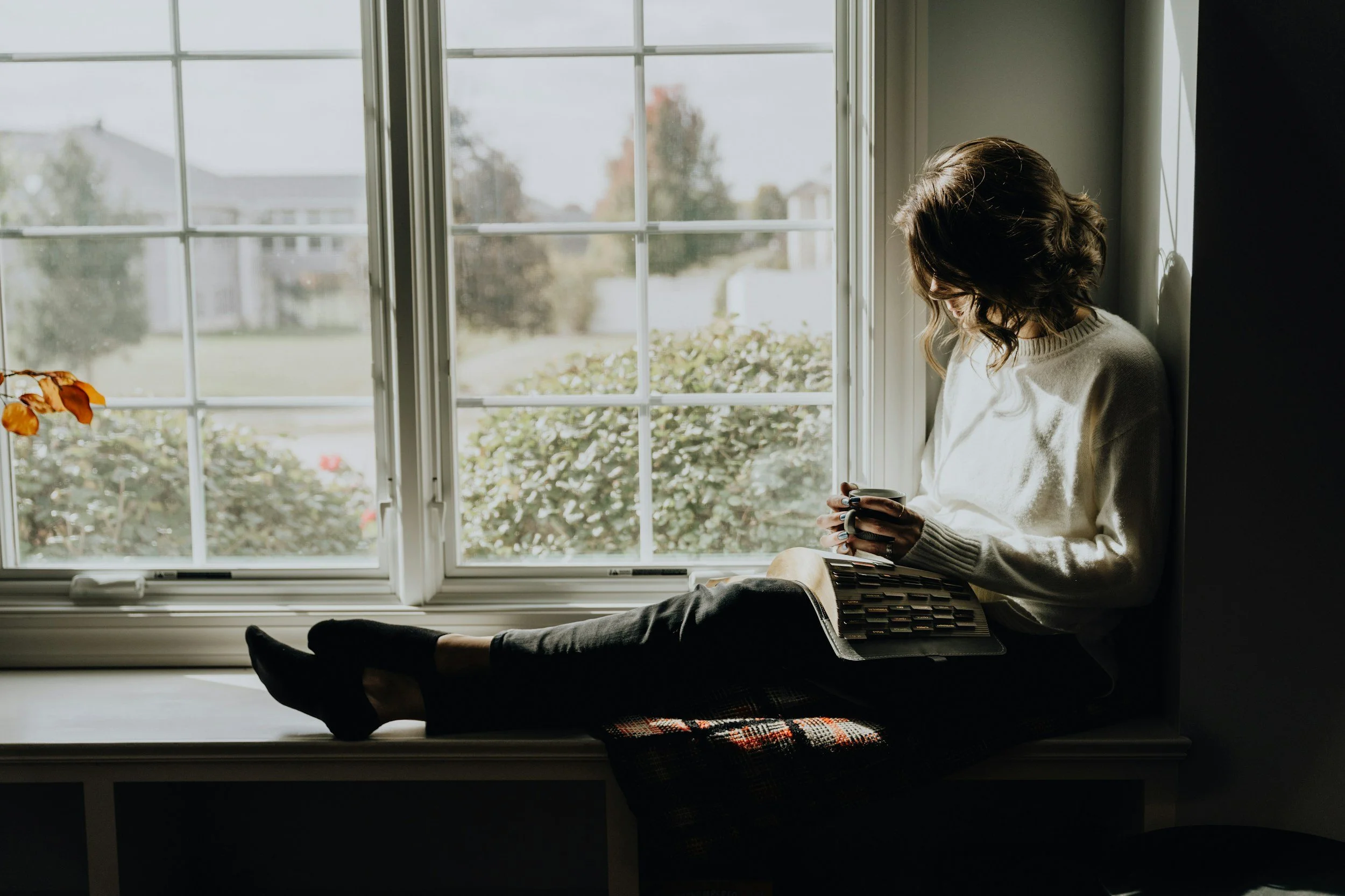 Woman reading a book by a window with coffee, relaxing in a cozy calm space — introvert self-care moment.