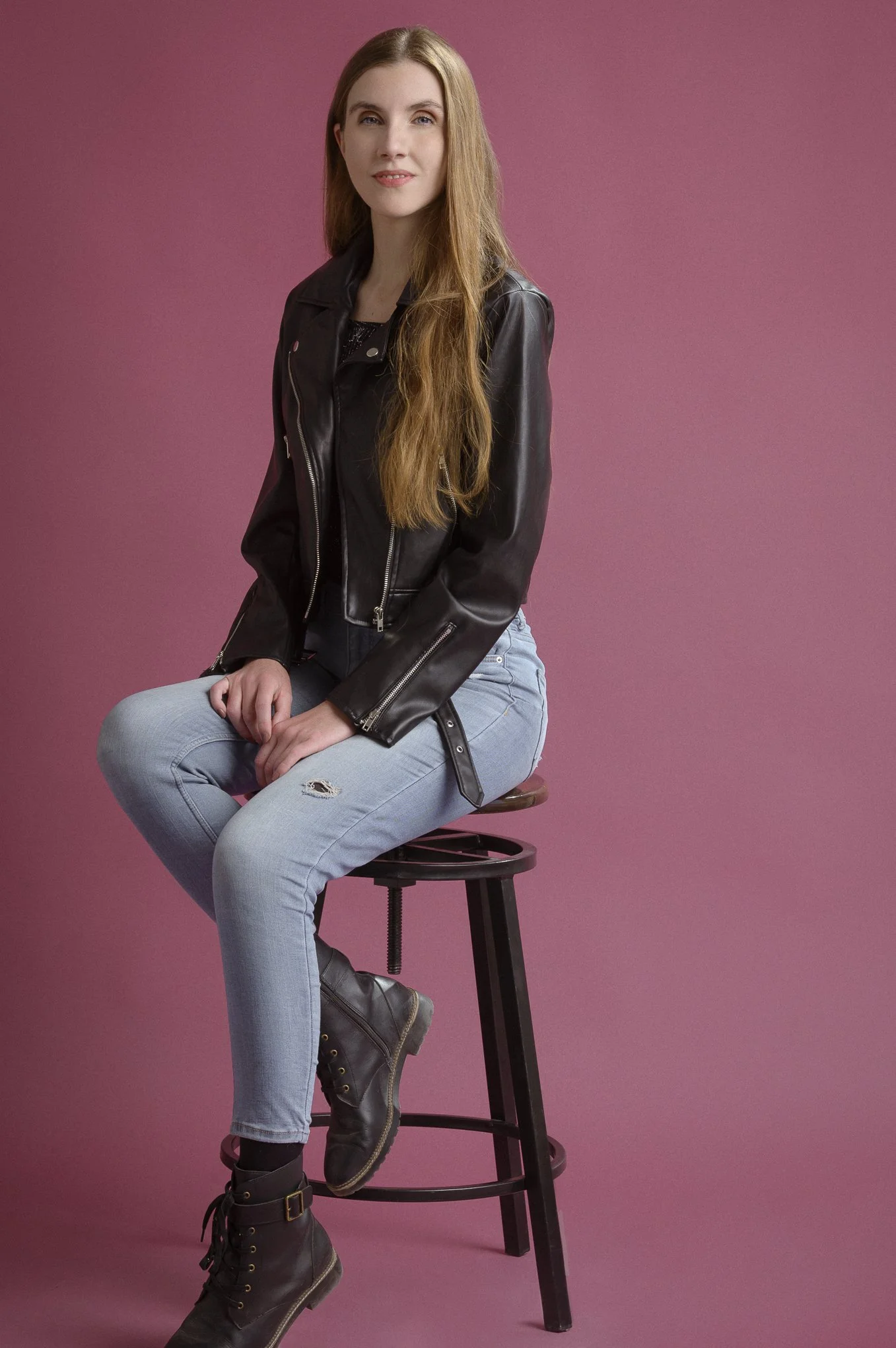 Ariane Vera sitting on a stool wearing jeans and a black leather jacket, smiling softly against a pink background.