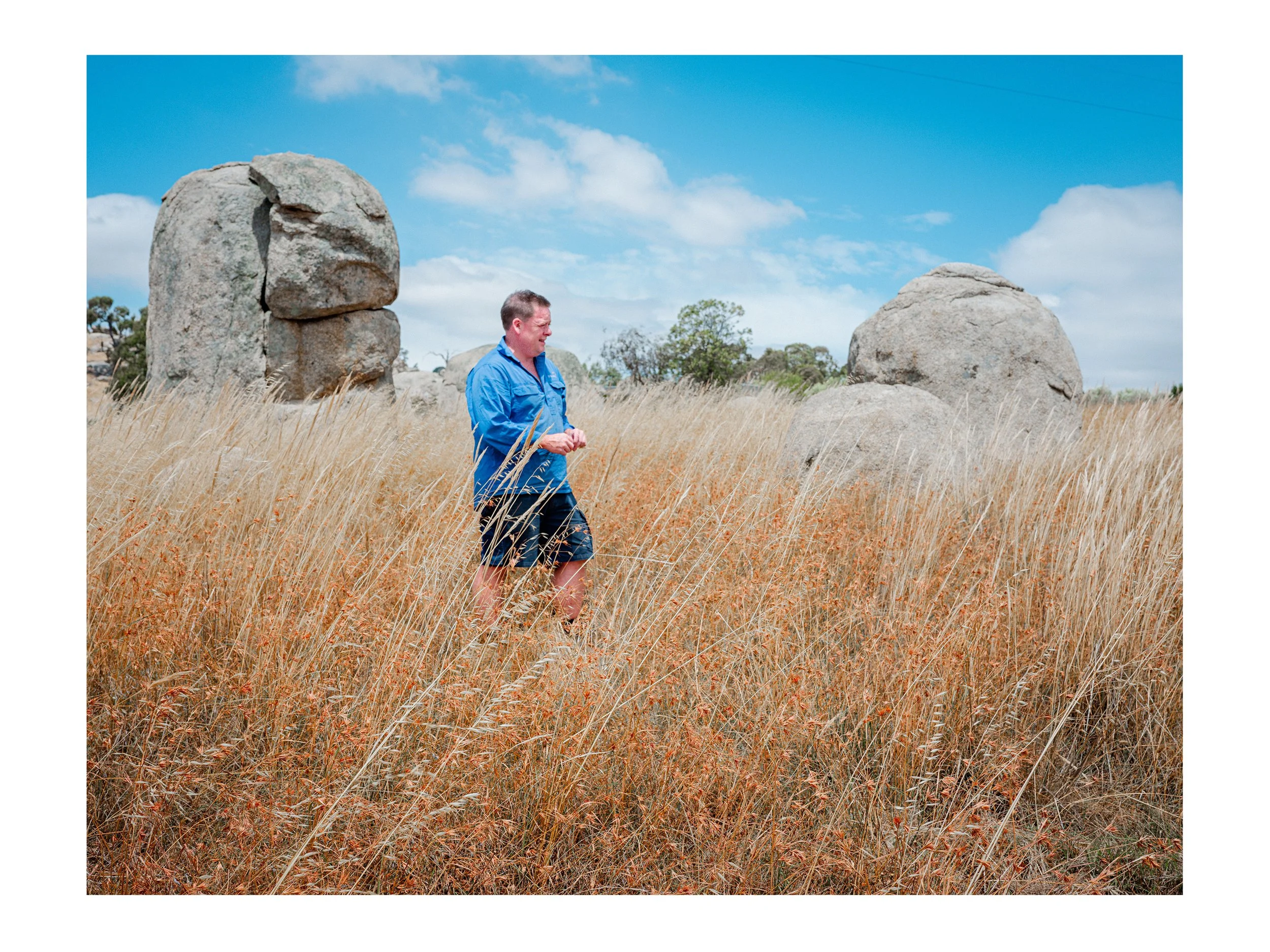 Winemaker Adam Foster of Syrahmi wines, walking through tall grass in a field with large rocks and a blue sky with some clouds.