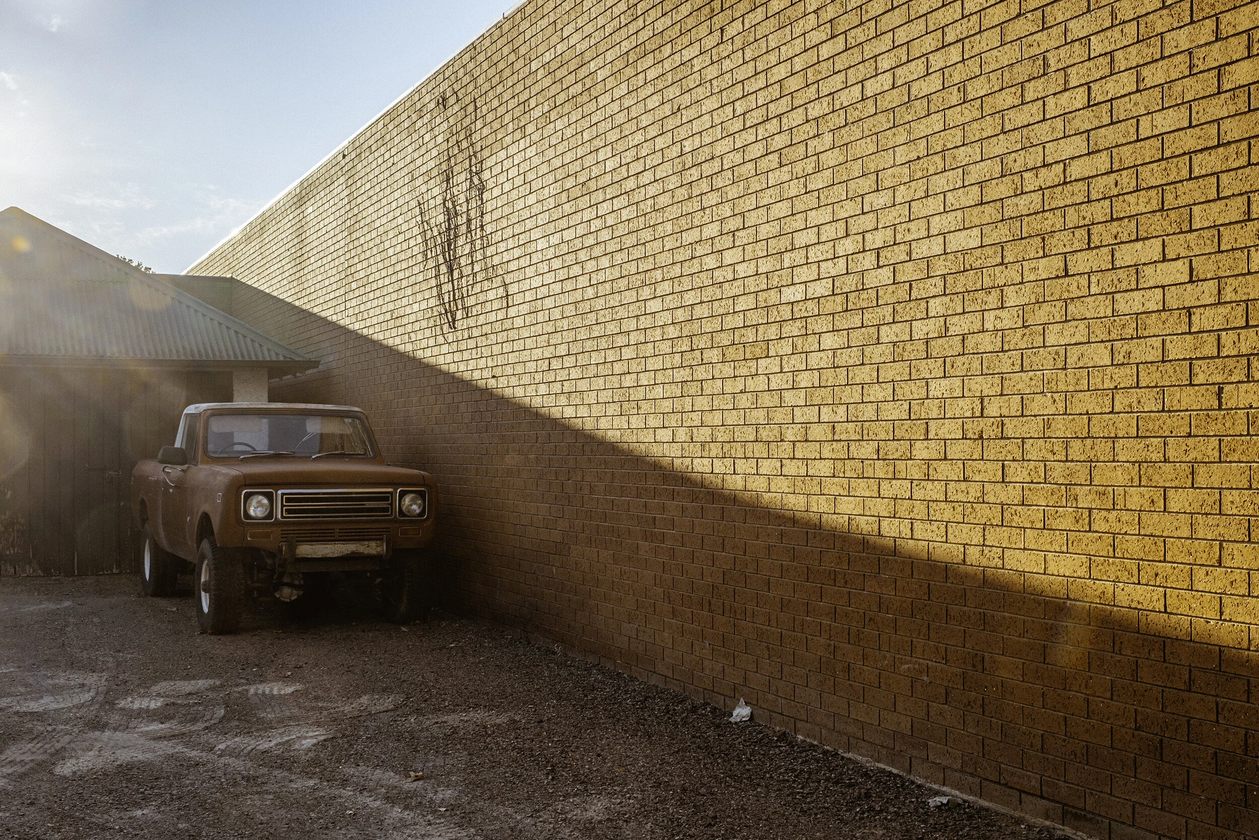 An old rust-colored truck parked next to a tall brick wall in Wangaratta with a leafless tree silhouette. The sun casts a shadow on the wall, with clear blue sky overhead.