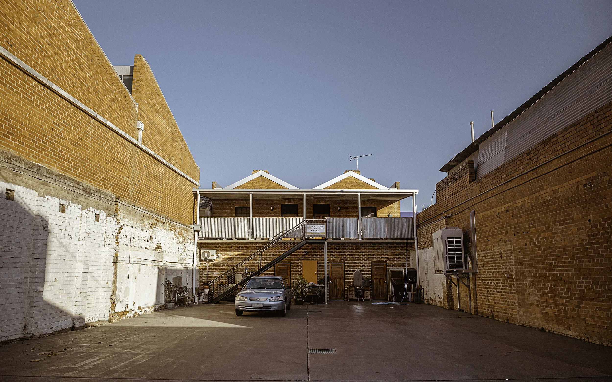 Brick building in Wangaratta and parking lot with a silver car, wooden doors, plants, chairs, air conditioning unit, and a metal staircase under a blue sky.