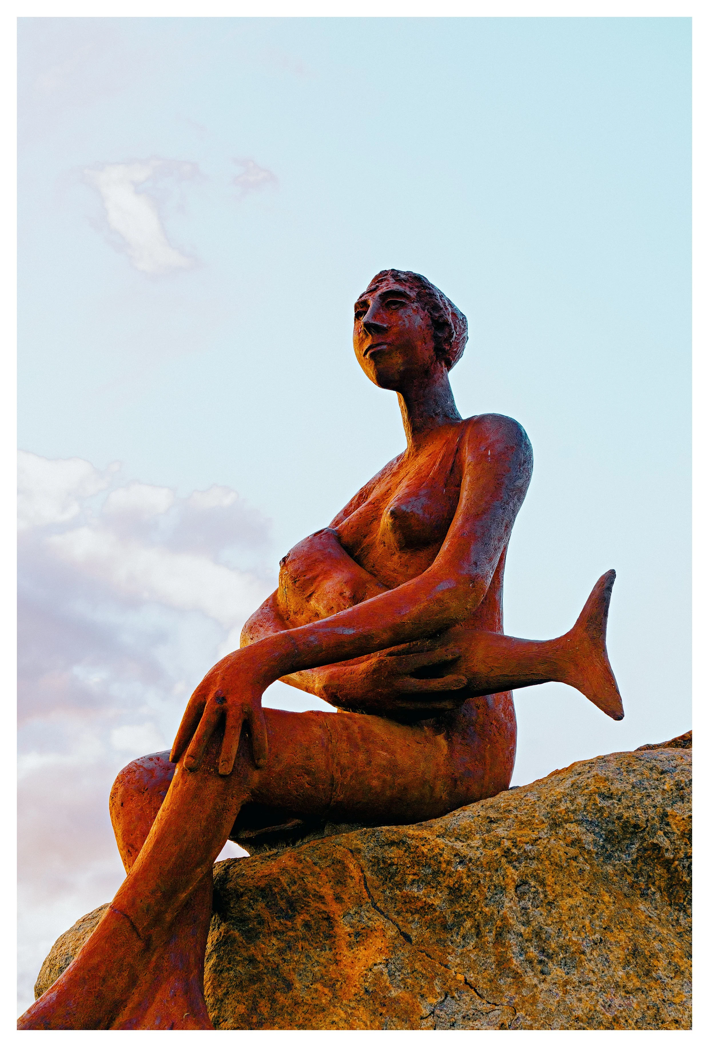 Statue of a woman on Margaret River beach with a fish in her lap, sitting on a large rock, against a sky with clouds.