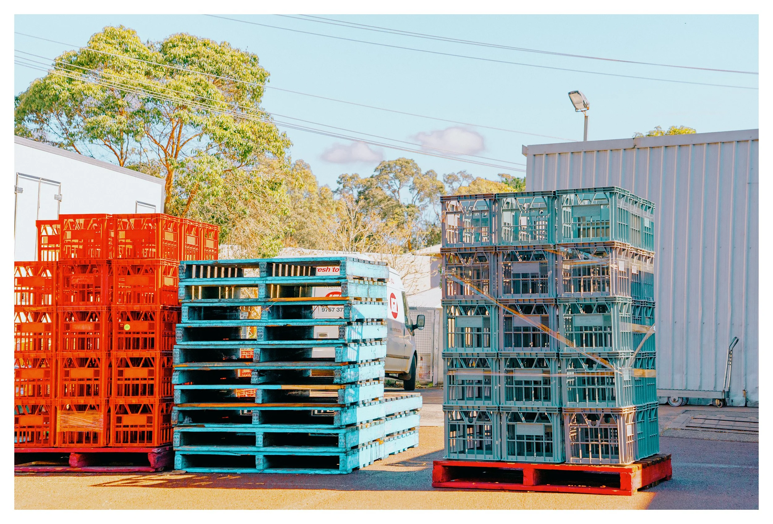 Stacked orange and teal plastic crates and pallets outside a Margaret River warehouse with trees and a blue sky in the background.