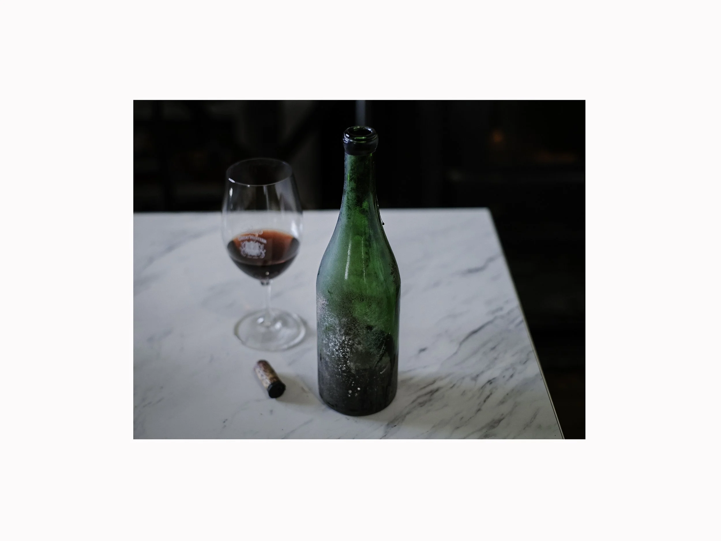A frosted green wine bottle, a glass of red wine, and a wine cork on a white marble table.