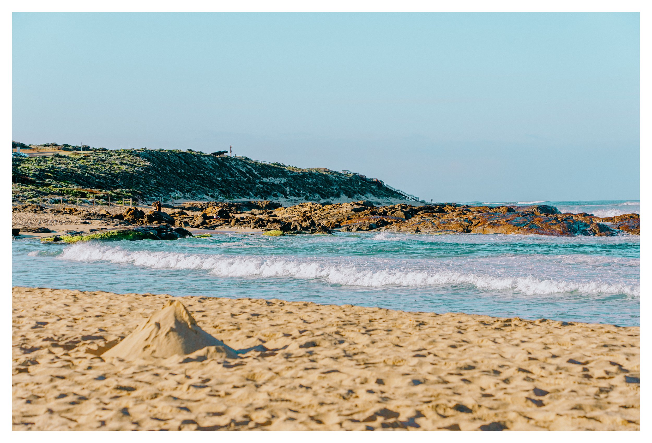 Sandy beach in Margaret River with a small sandcastle, gentle waves, rocky shoreline, and a hillside in the distance on a clear day.