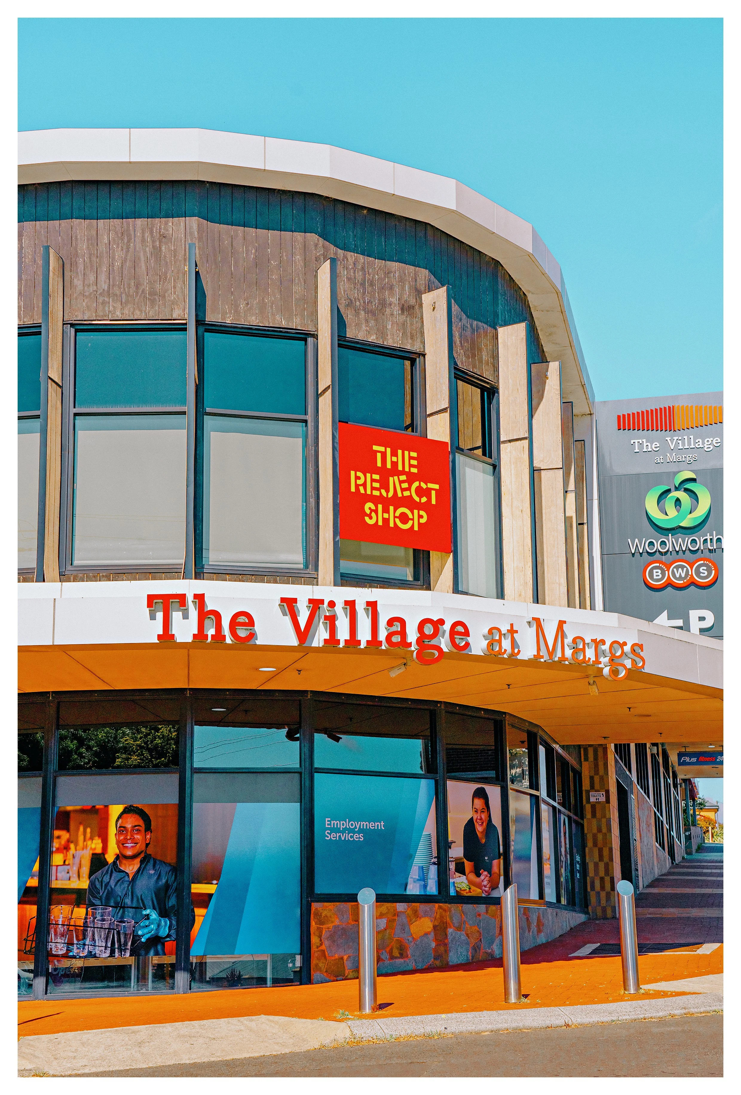 A shopping center building in Margaret River with signs for The Village at Margs and Woolworth. The building has large windows, a curved roof, and a sign for The Reject Shop.