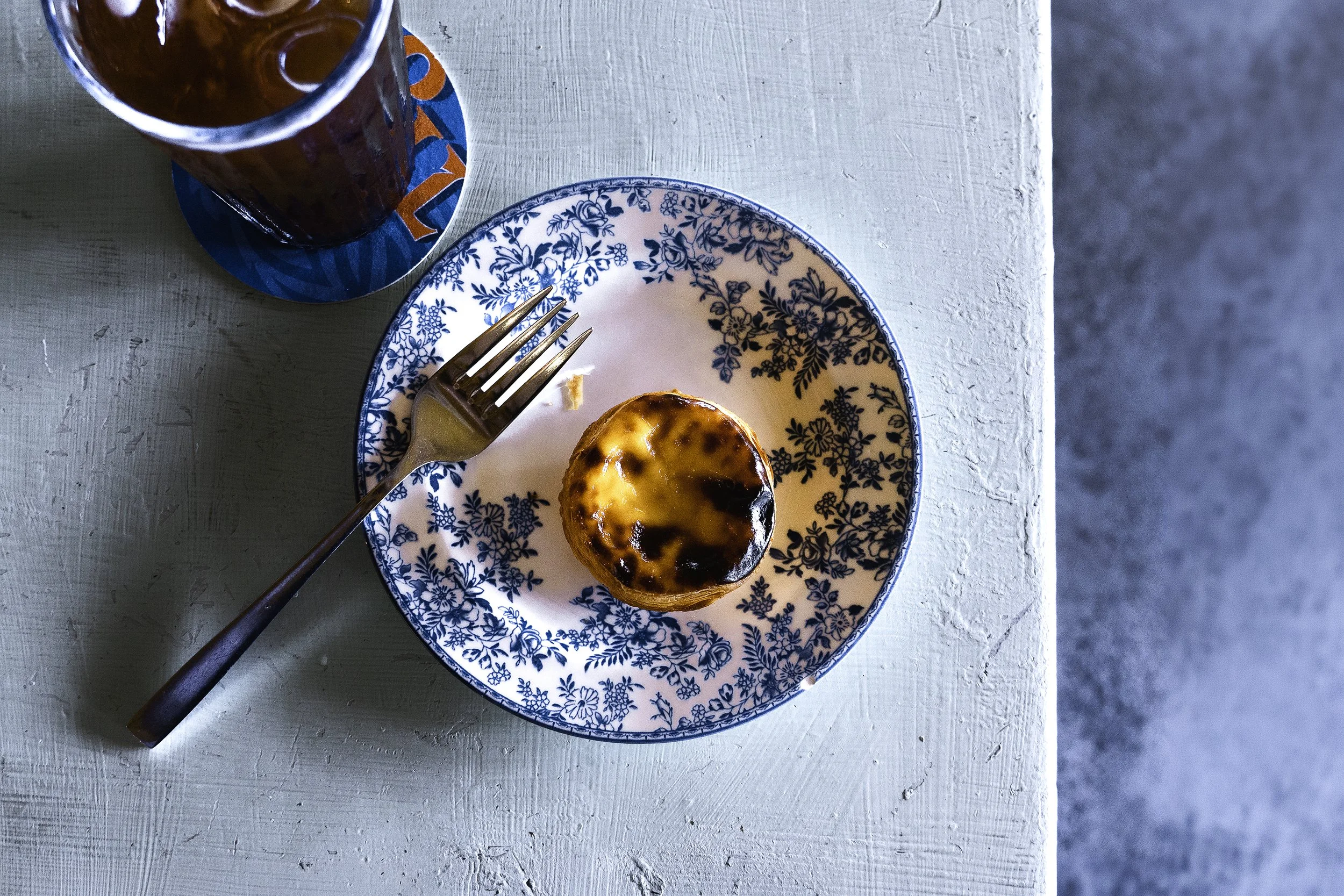 A single pastel de nata Portuguese tart sits on a blue plate, on a blue table, at the Bartolo cafe in Kuala Lumpur.