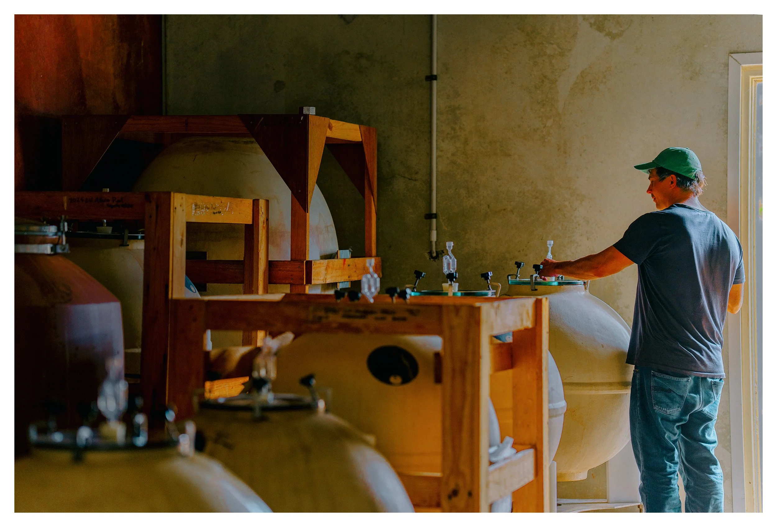 Winemaker Nic Peterkin wearing a green cap and a dark t-shirt is working with large fermentation tanks in a winery or brewery.