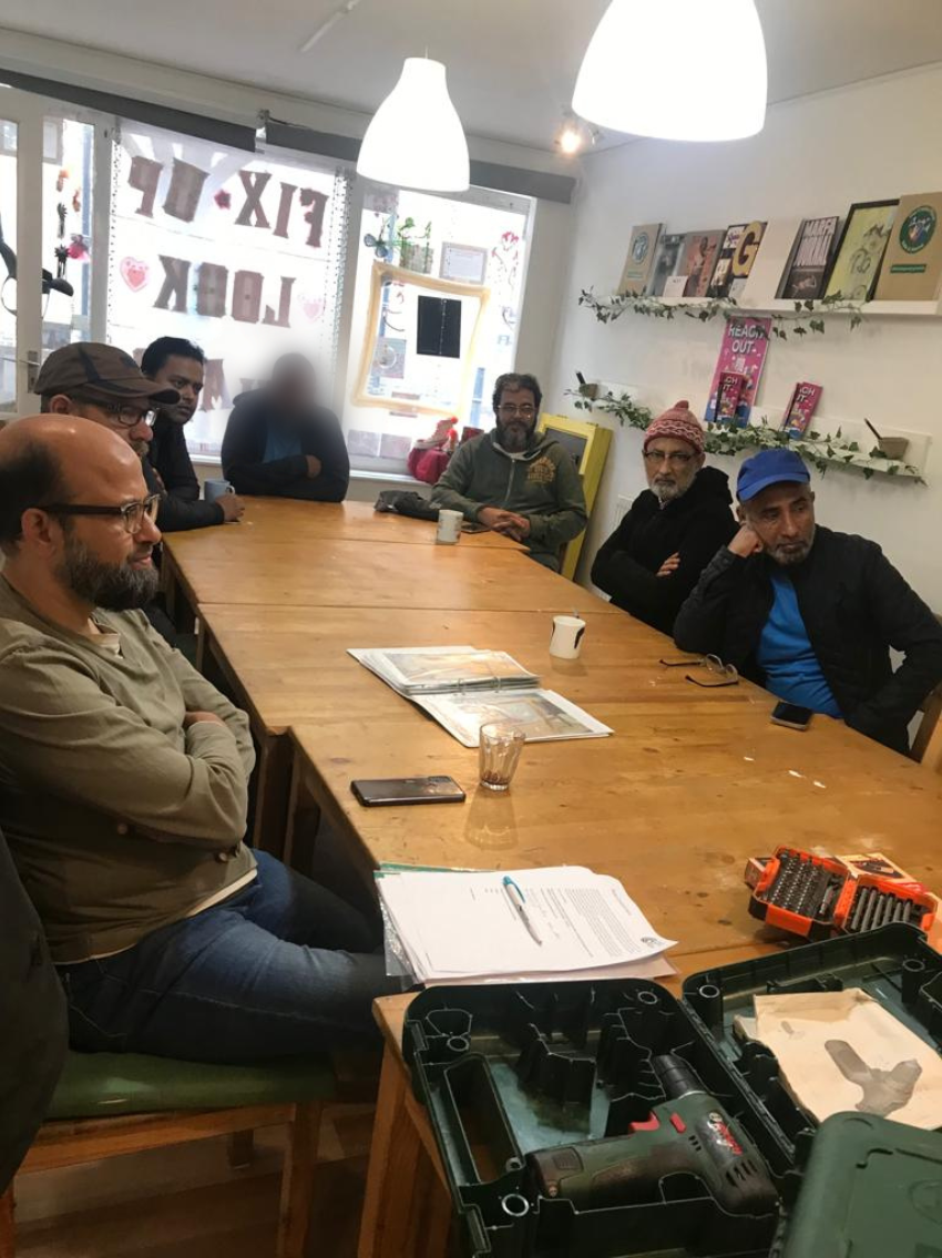 Group_of_men_at_table_with_tools_in_foreground.png