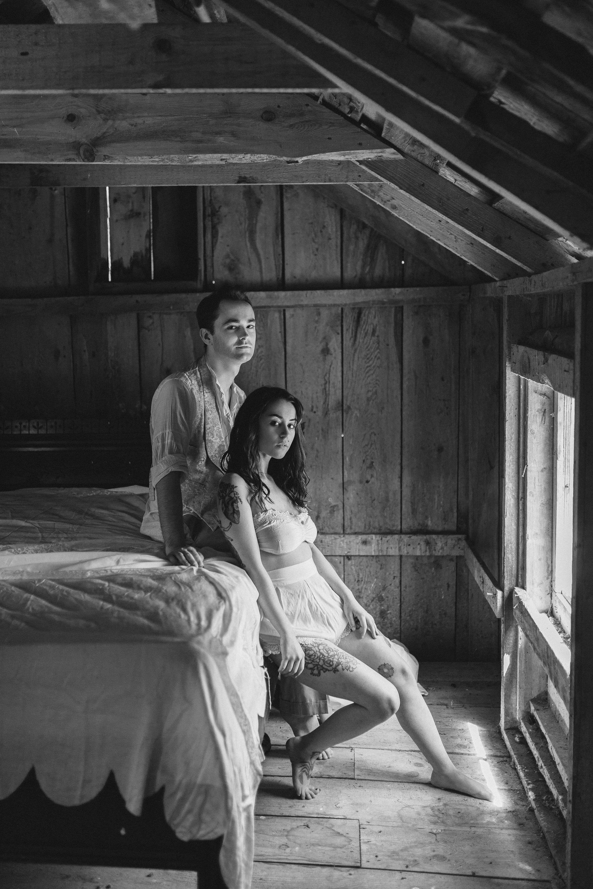 Black and white photograph of two women in a rustic wooden room with a sloped ceiling. One woman with dark hair and tattoos sits on a bed, her right leg extended and foot on the floor, while the other woman with dark hair tied back stands behind her,