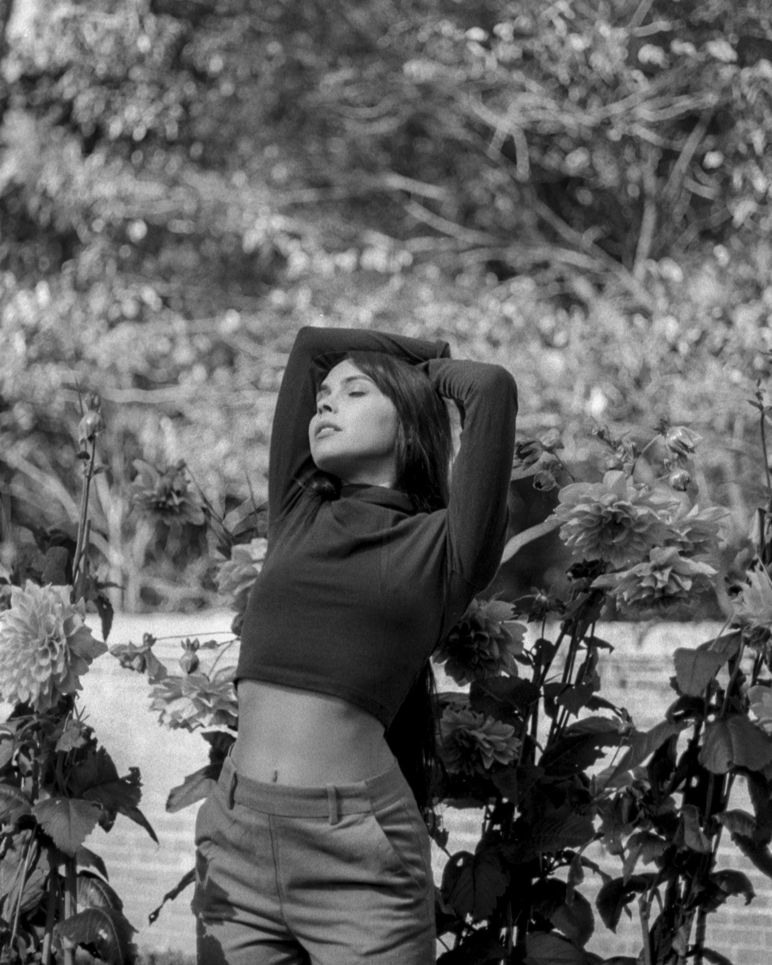 A woman with long hair posing with her eyes closed, wearing a dark jacket and light pants, surrounded by large flowers and foliage, in an outdoor setting.