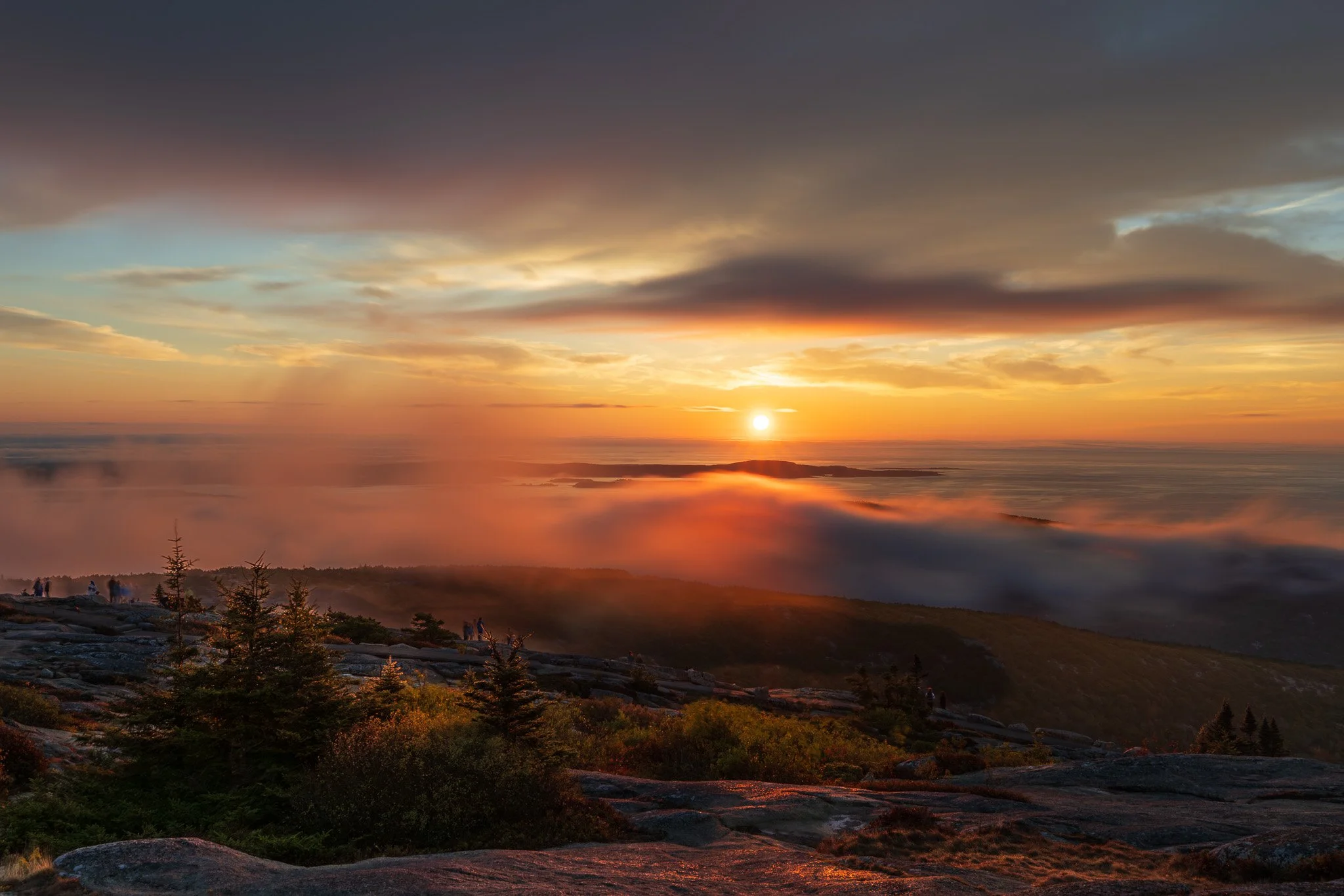 Sunset over a foggy mountain landscape with trees and rocks in the foreground.