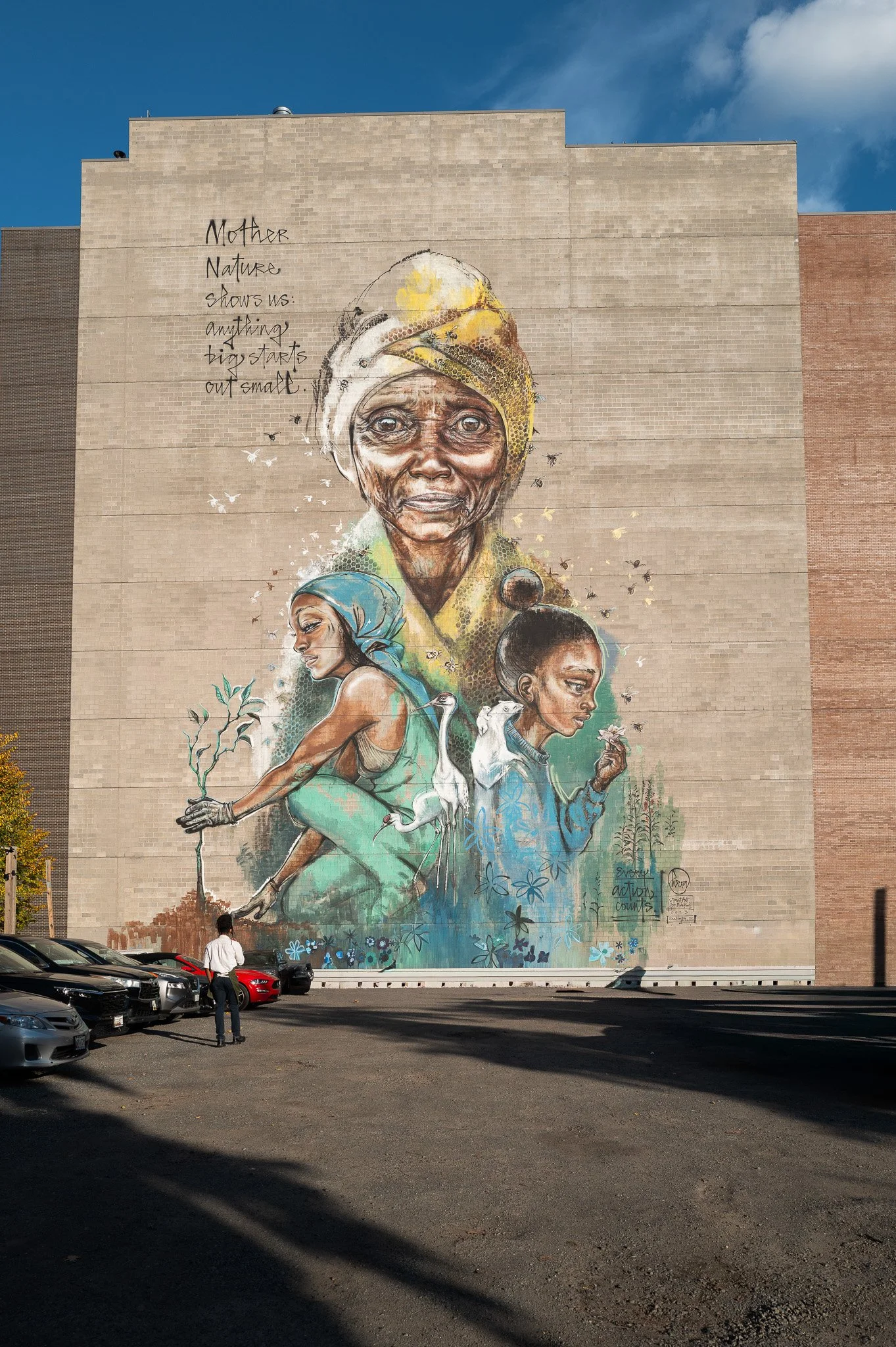 A large mural on the side of a building depicting an elderly woman with a head wrap, with two young women and a crane, surrounded by a nature scene and birds. The mural includes the quote, "Mother nature shows us: anything big starts out small."