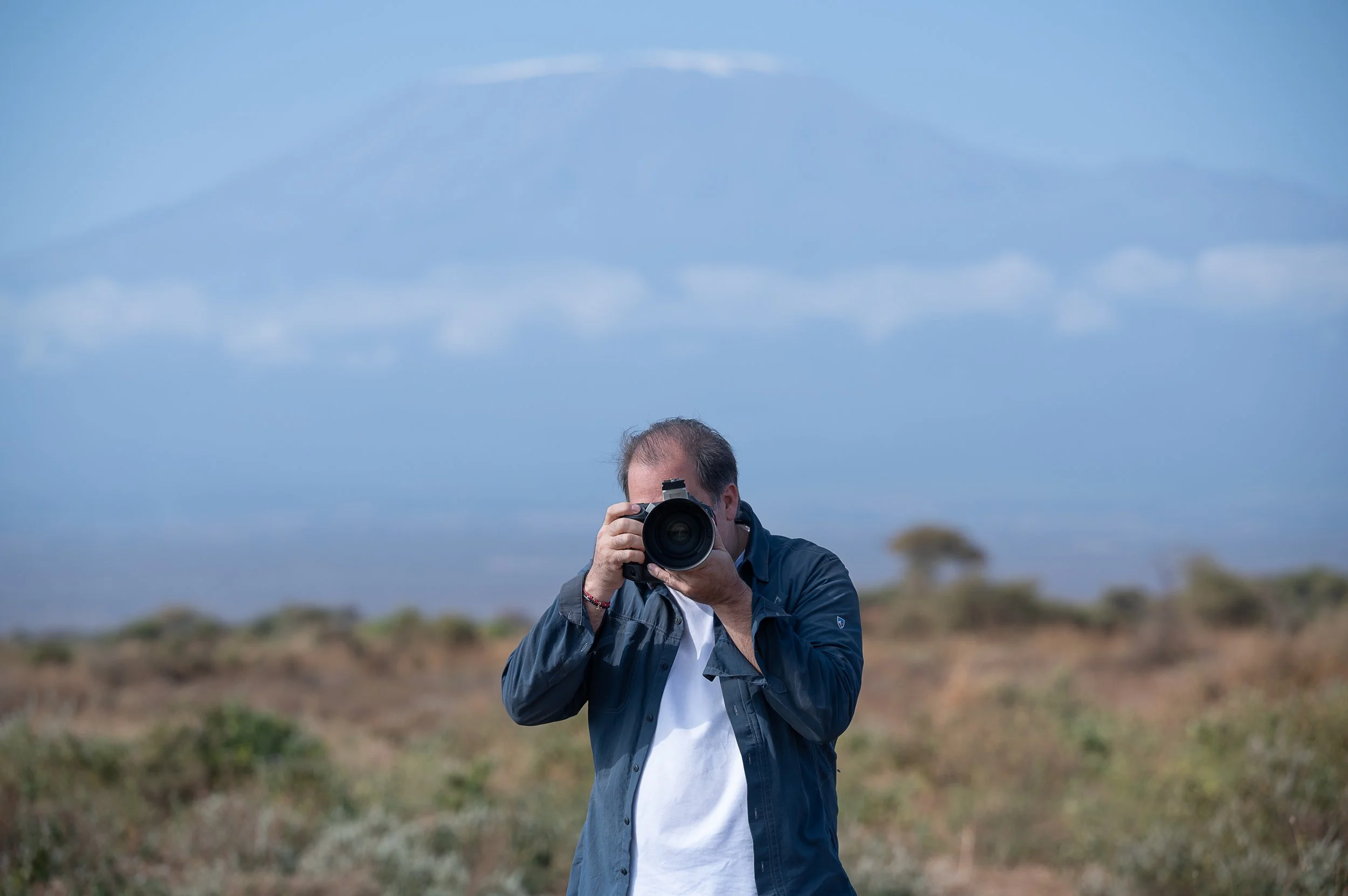 Man taking a photograph outdoors in a field with a camera, with a mountain and partly cloudy sky in the background.