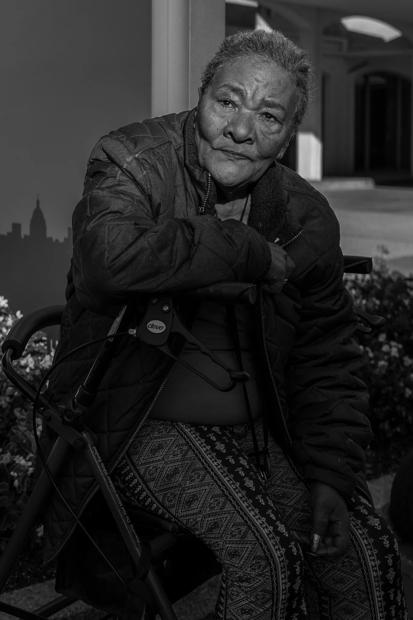 An elderly woman sitting on a walker with a cityscape silhouette in the background, in black and white.