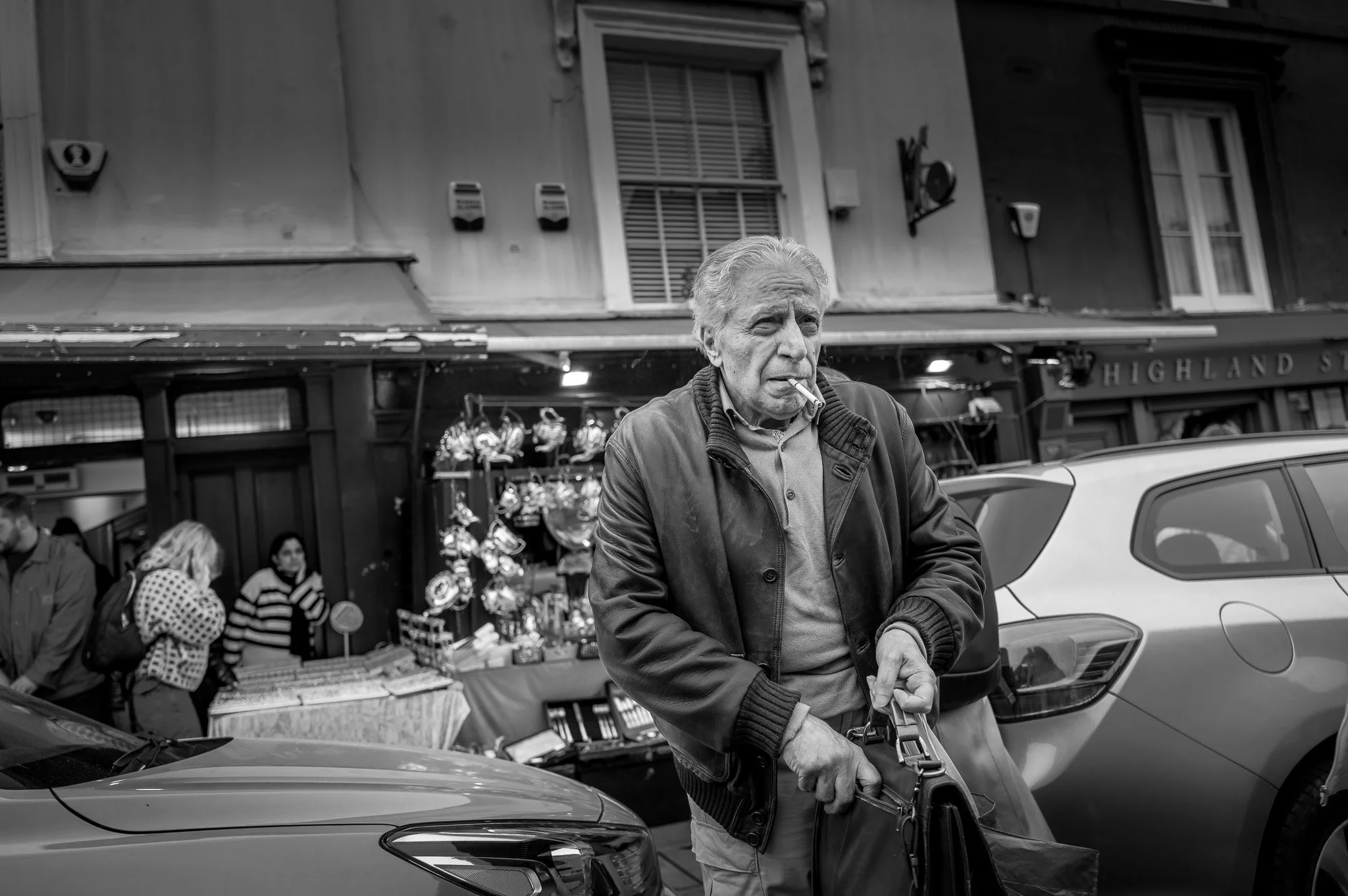 An elderly man with gray hair and a cigarette in his mouth walks on a city street, carrying a bag, with people and shops in the background.