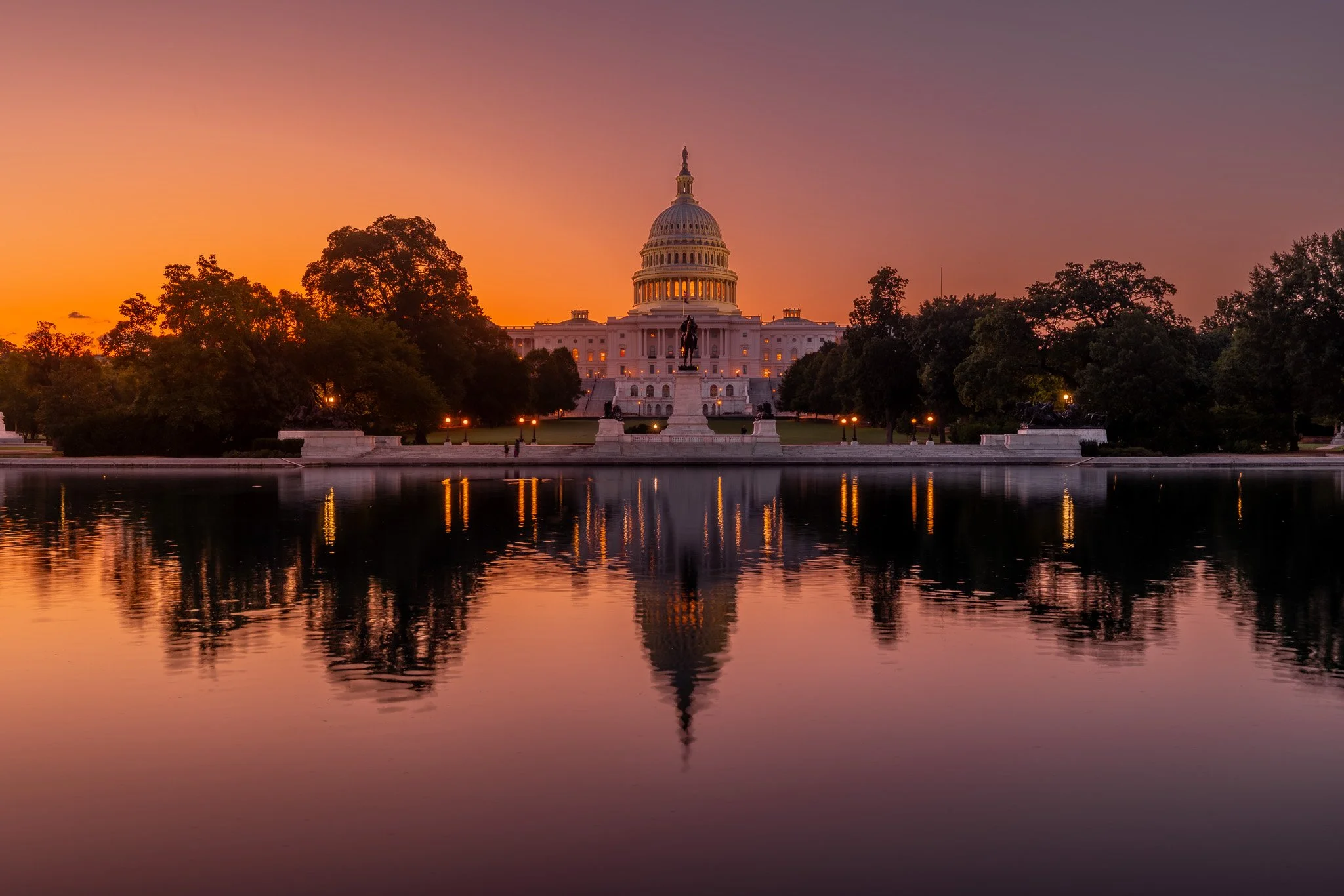 The United States Capitol building at sunset, with sunset sky, trees, and water reflecting the scene.