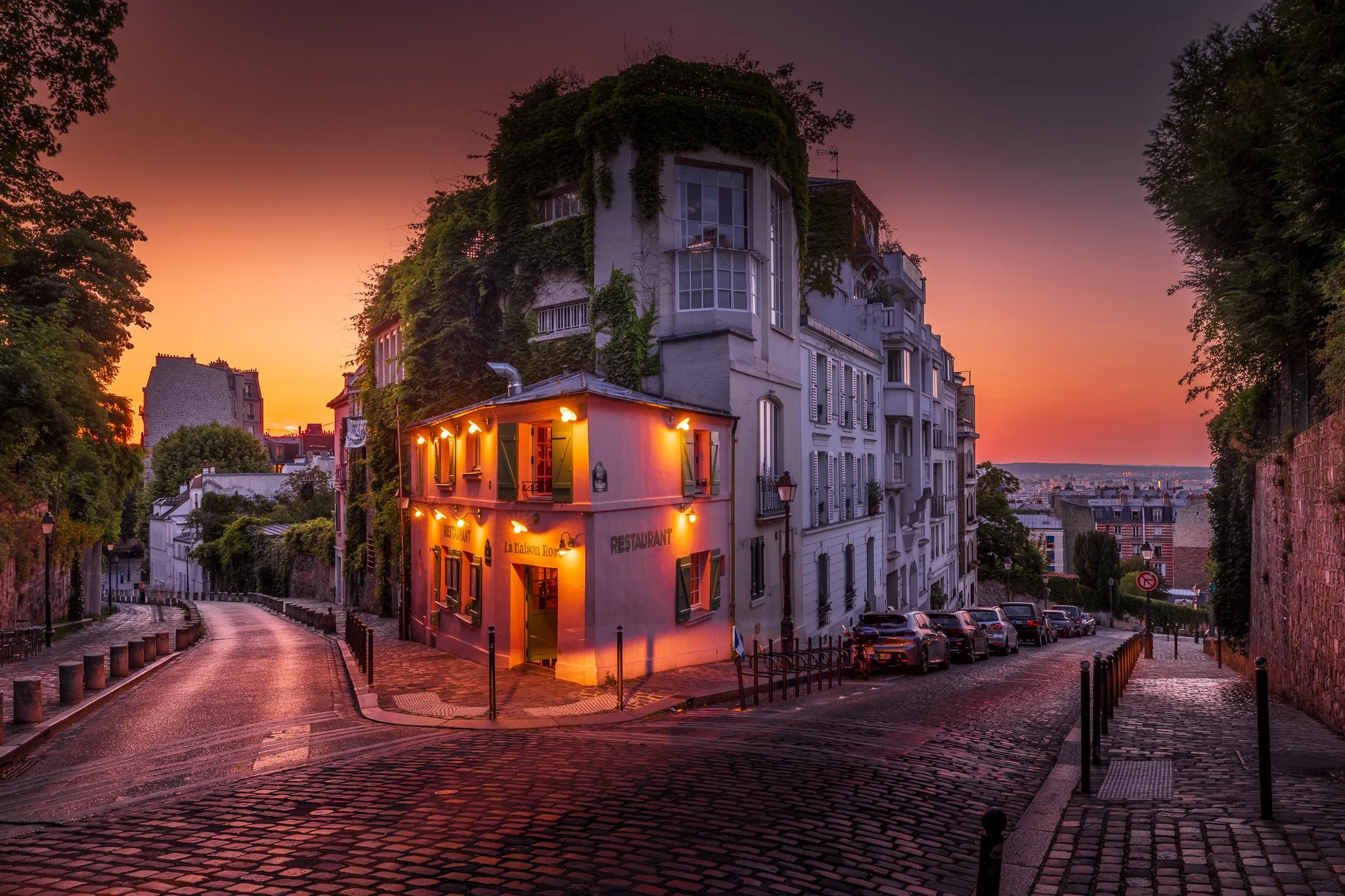 A cobblestone street in a European city at sunset, featuring a small yellow restaurant with warm lighting, surrounded by tall white buildings covered in greenery.
