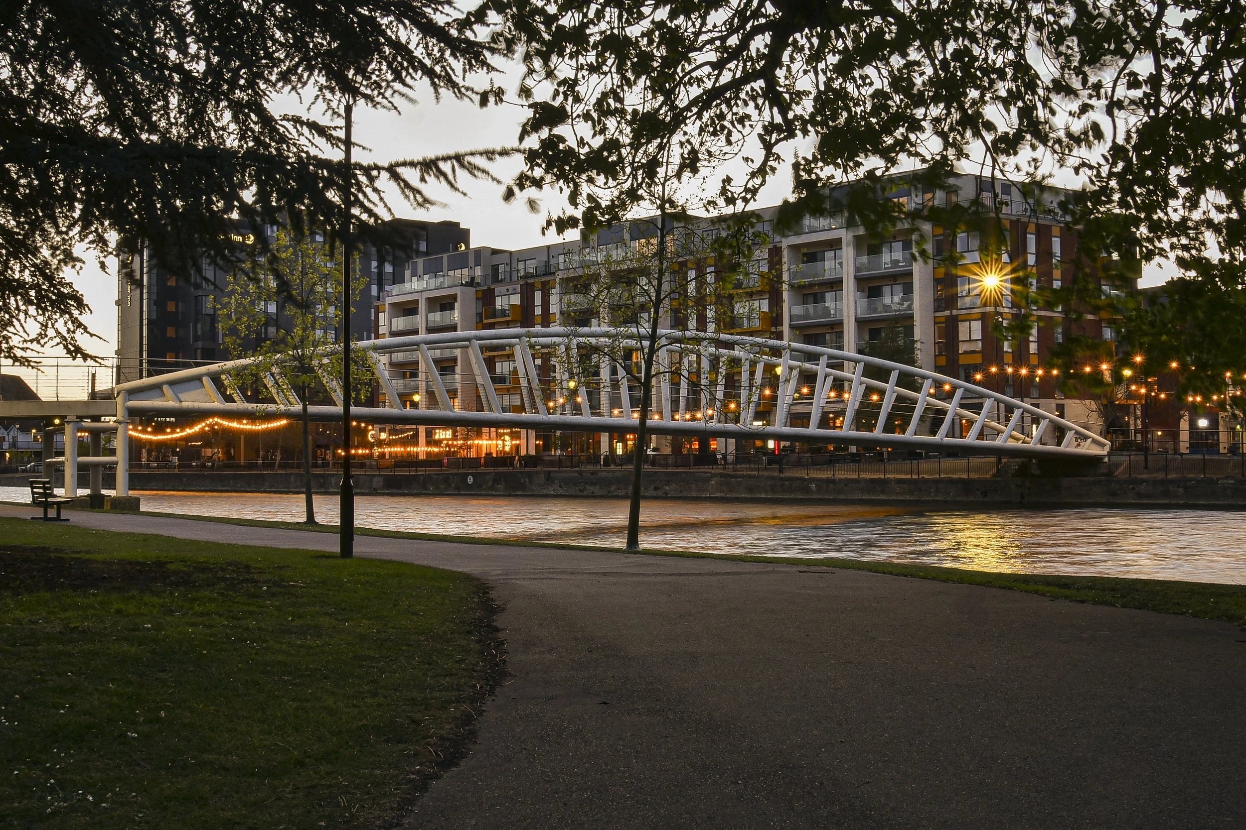 A modern white pedestrian bridge over a river at dusk, with colorful apartment buildings and streetlights in the background, framed by trees in the foreground.